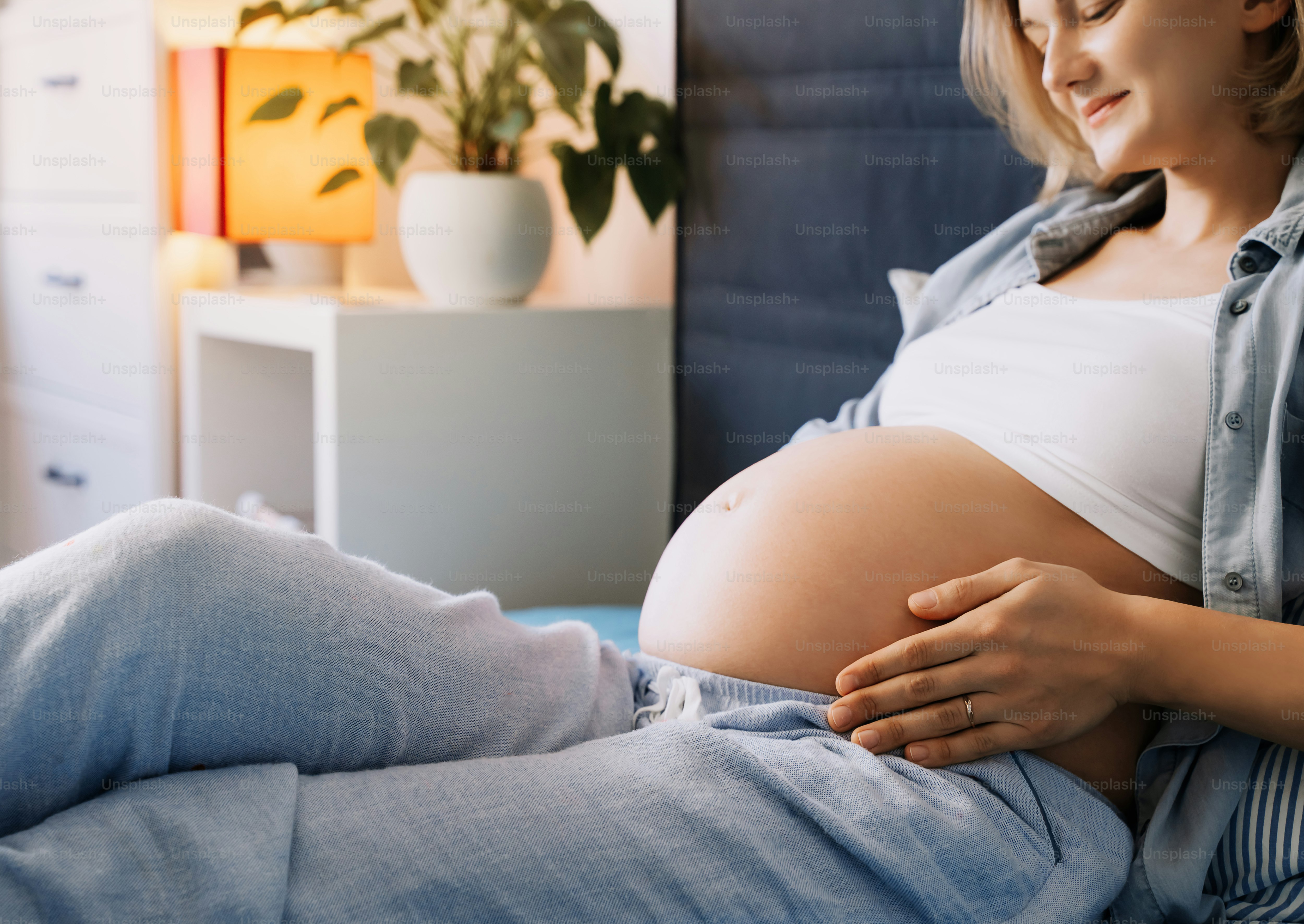 A pregnant woman in a white top and blue pants resting on a bed
