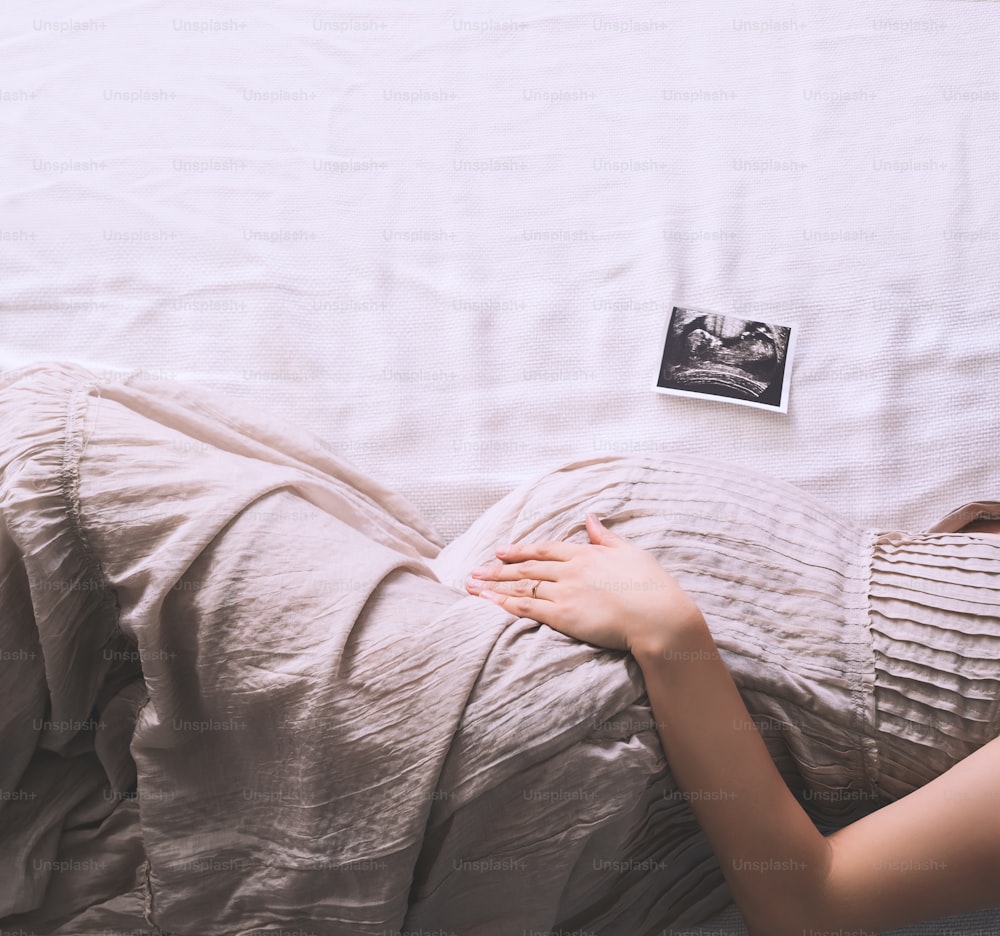 Beautiful pregnant woman in dress holding hands on belly lying on side in profile in bed. Expectant mother waiting and preparing for baby birth. Pregnancy photo with happy and gentle mood.