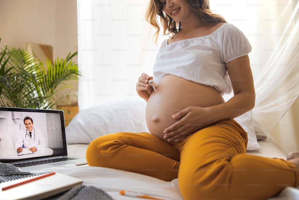 Beautiful expectant mother sitting on bed and having video online consultation with doctor stock photo