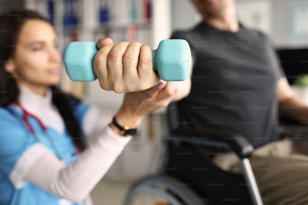 Girl doctor helps patient train with dumbbell. Handicapped man sits working out dumbbell arm. Treatment and restoration lost functions. Effective exercises to restore physical activity hand