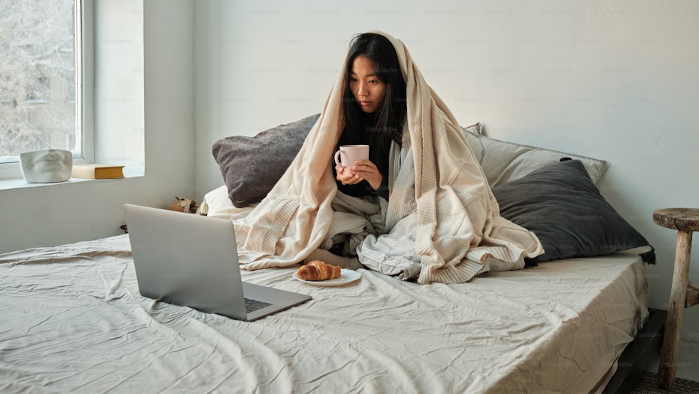 uma mulher sentada em uma cama com um laptop e um cobertor sobre a cabeça