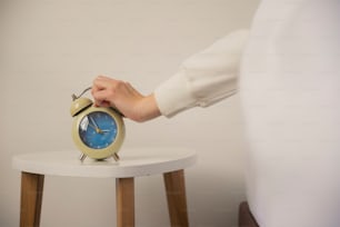 uma pessoa segurando um despertador em uma pequena mesa