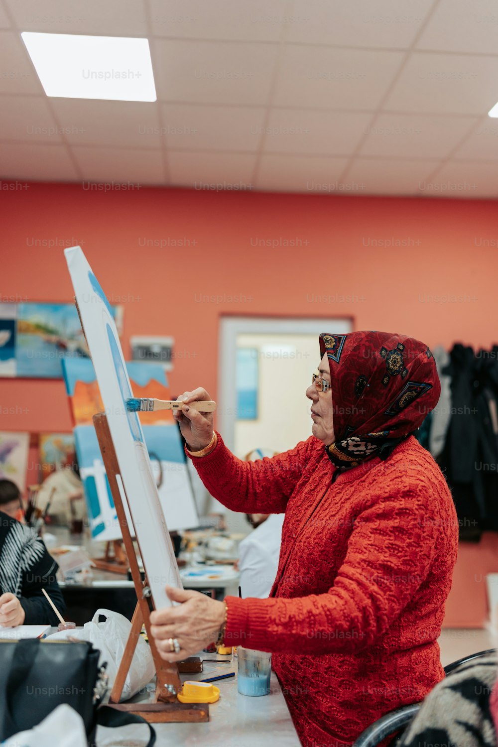 a woman in a red jacket is painting on a easel