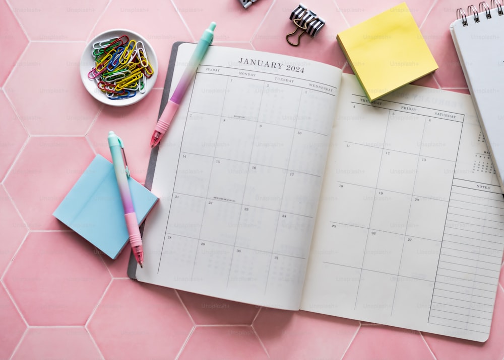un calendario, bolígrafos, marcadores y otros suministros de oficina sobre una superficie rosa
