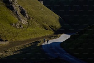 a couple of people walking down a road next to a lush green hillside