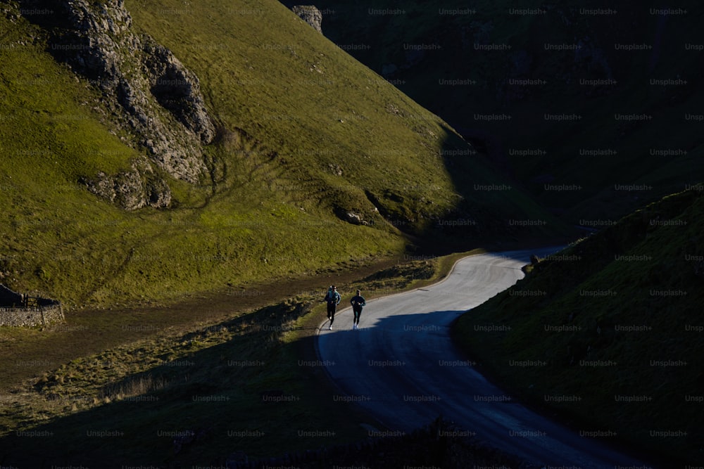 a couple of people walking down a road next to a lush green hillside