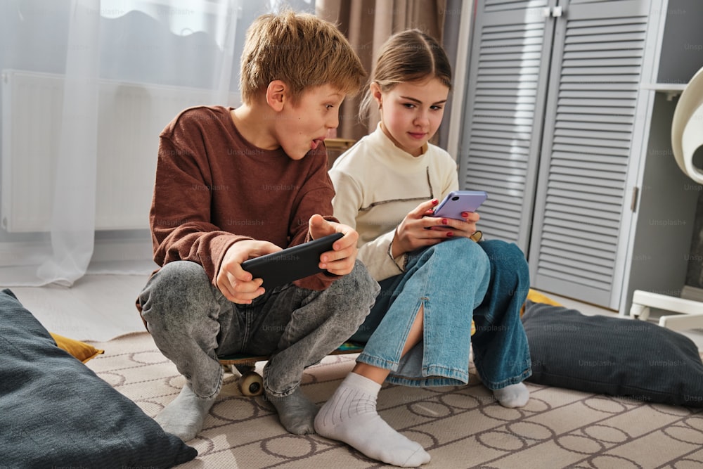 a boy and a girl sitting on the floor looking at a cell phone