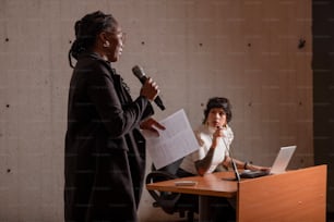 a woman speaking into a microphone next to a woman sitting at a desk