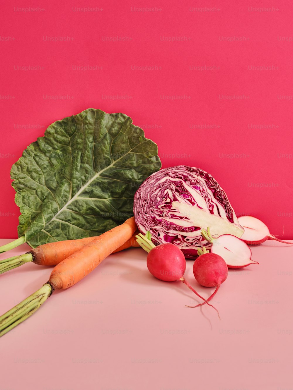 a cabbage, radishes, and carrots on a pink surface