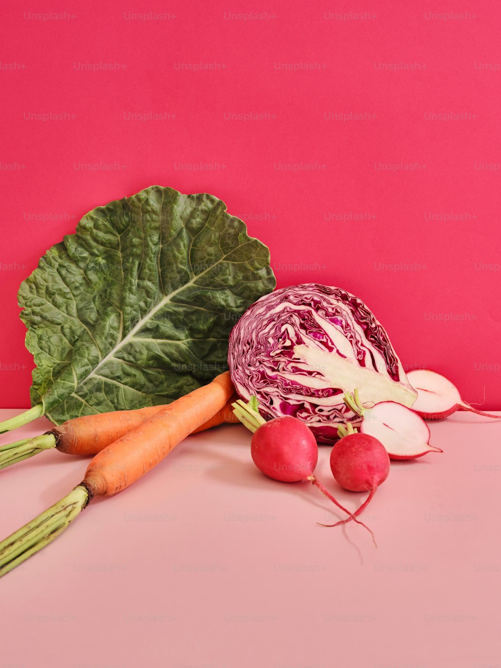 um repolho, rabanetes e cenouras em uma superfície rosa