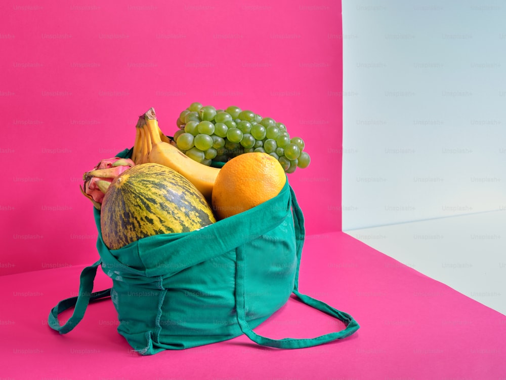 una bolsa verde llena de fruta sobre un fondo rosa