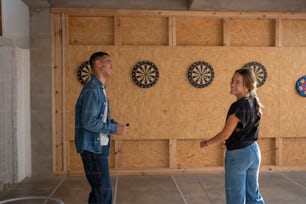 a man and a woman standing in a room with darts on the wall