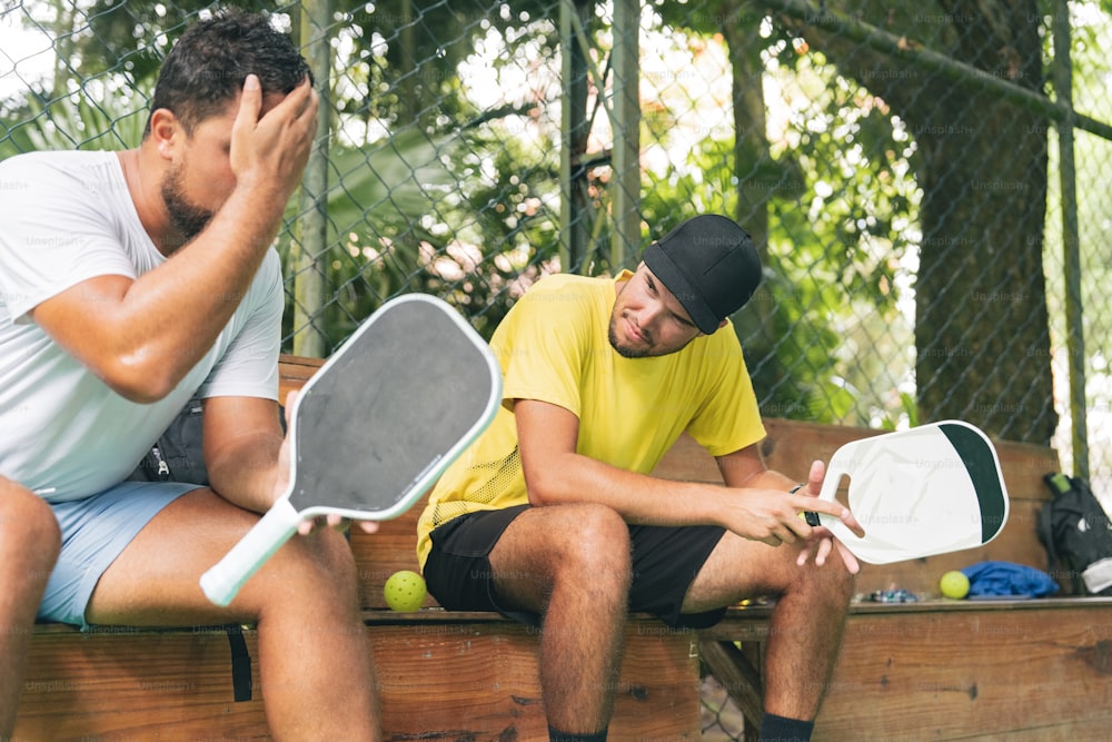 two men sitting on a bench holding tennis rackets