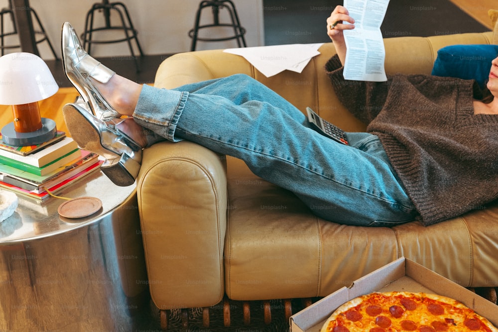 uma mulher sentada em um sofá lendo um papel e comendo uma pizza