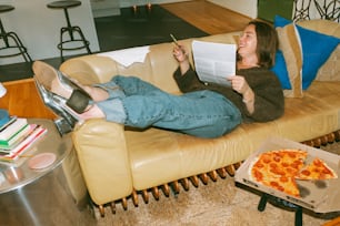 a woman sitting on a couch reading a book and eating pizza