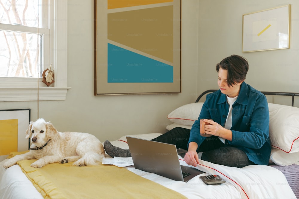 a woman sitting on a bed with a laptop and a dog