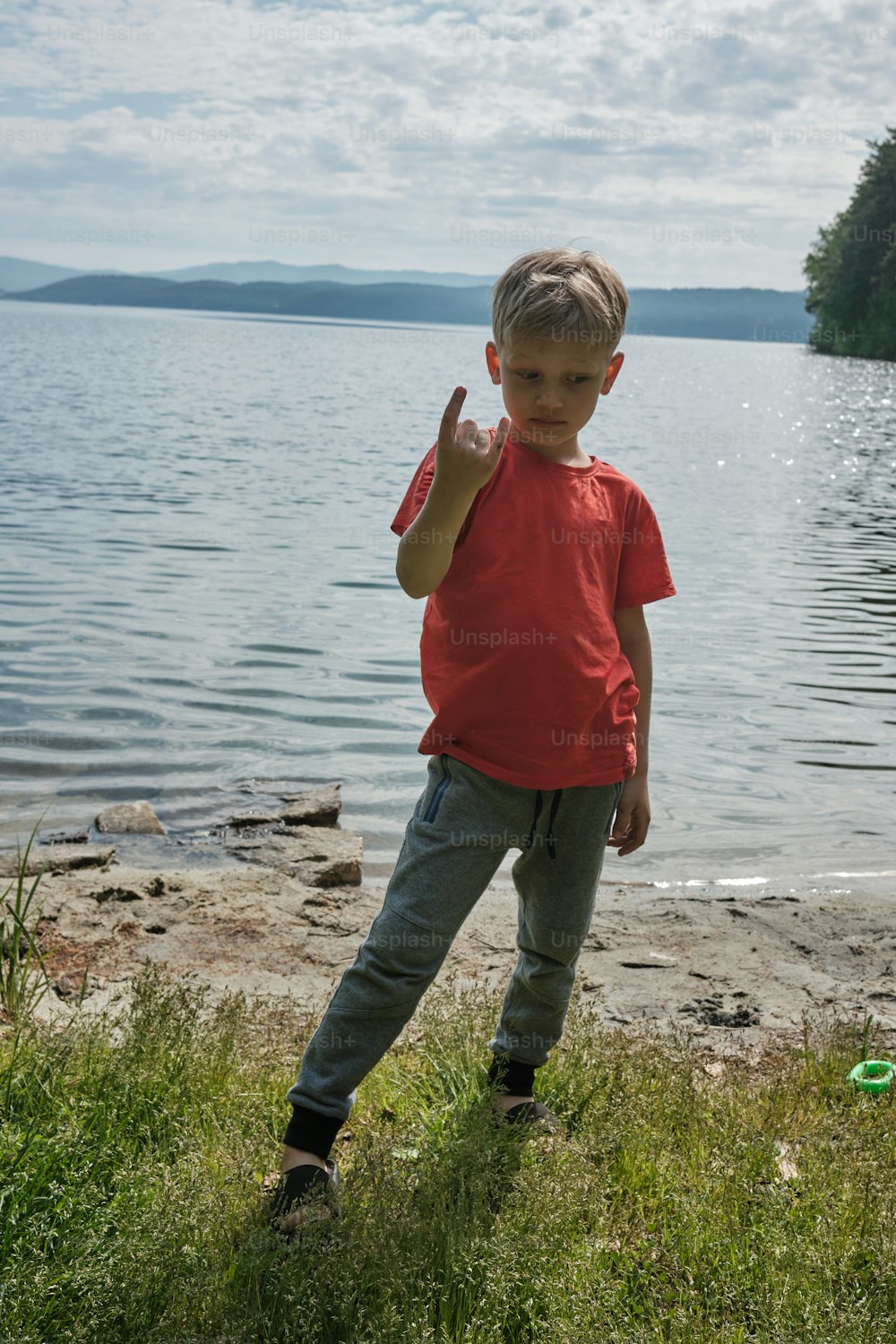 a young boy standing on the shore of a lake giving the peace sign