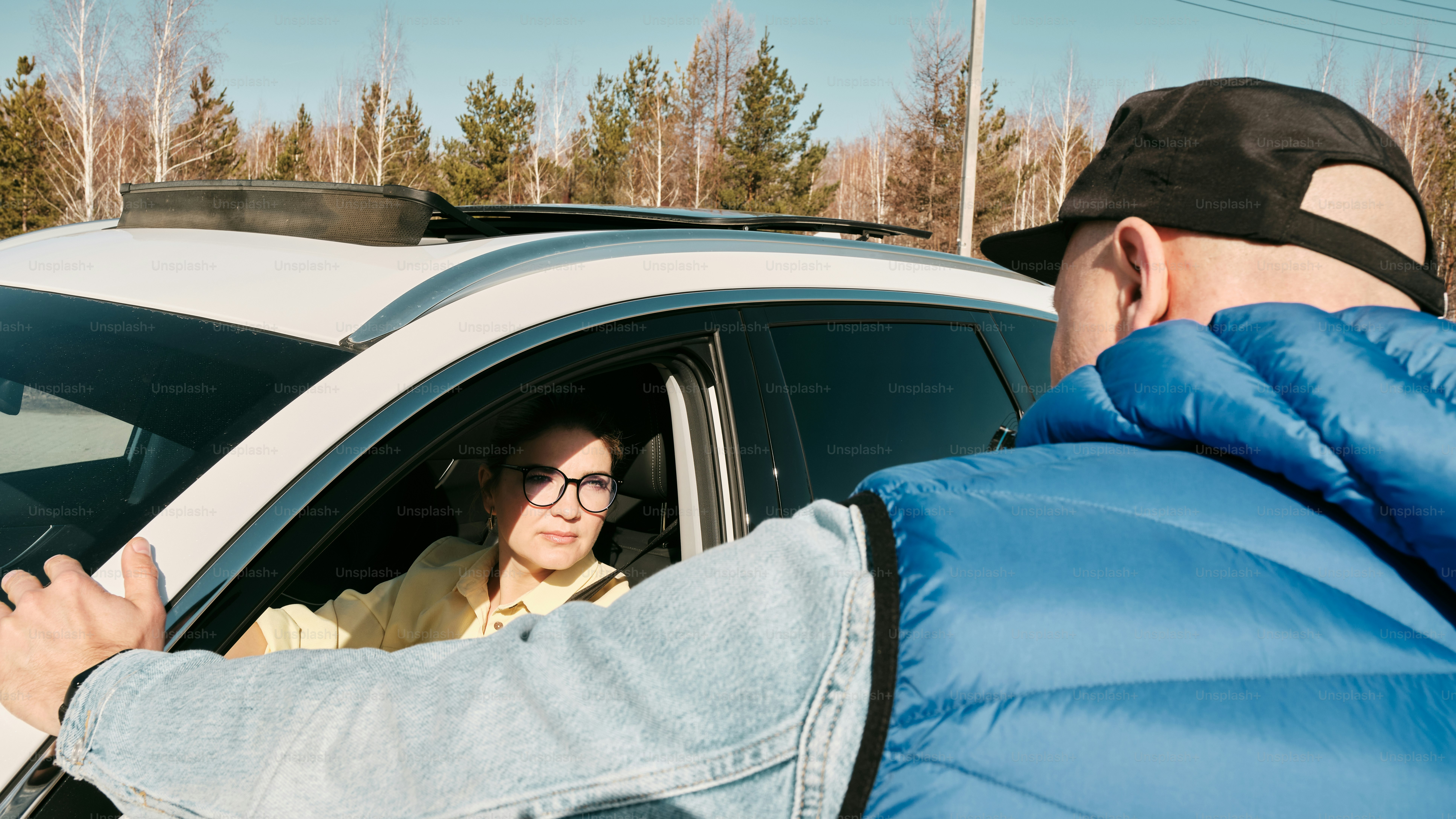 Un hombre con una chaqueta azul acolchada hablando con una mujer en un coche blanco