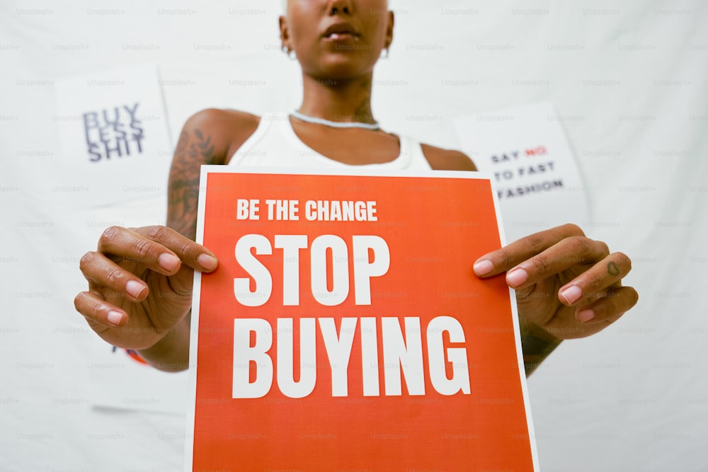 a woman holding a sign that says be the change stop buying