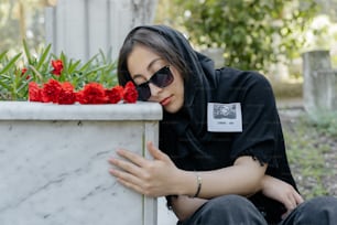uma mulher com capuz preto e óculos escuros encostada a uma floreira de mármore
