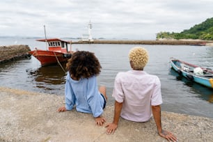 a man and a woman sitting on the edge of a pier looking at a boat