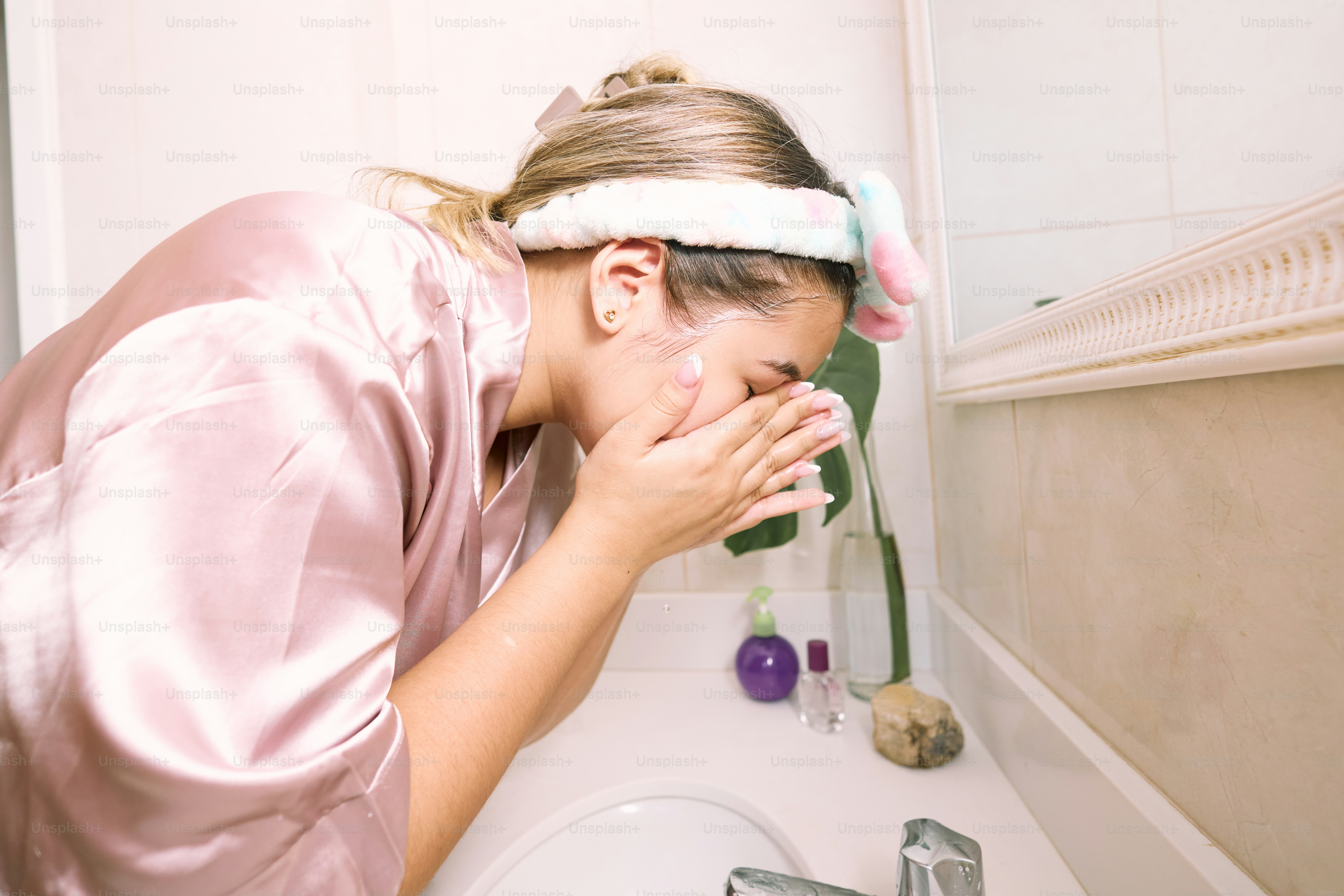 a woman in a pink robe wiping her face in front of a bathroom sink