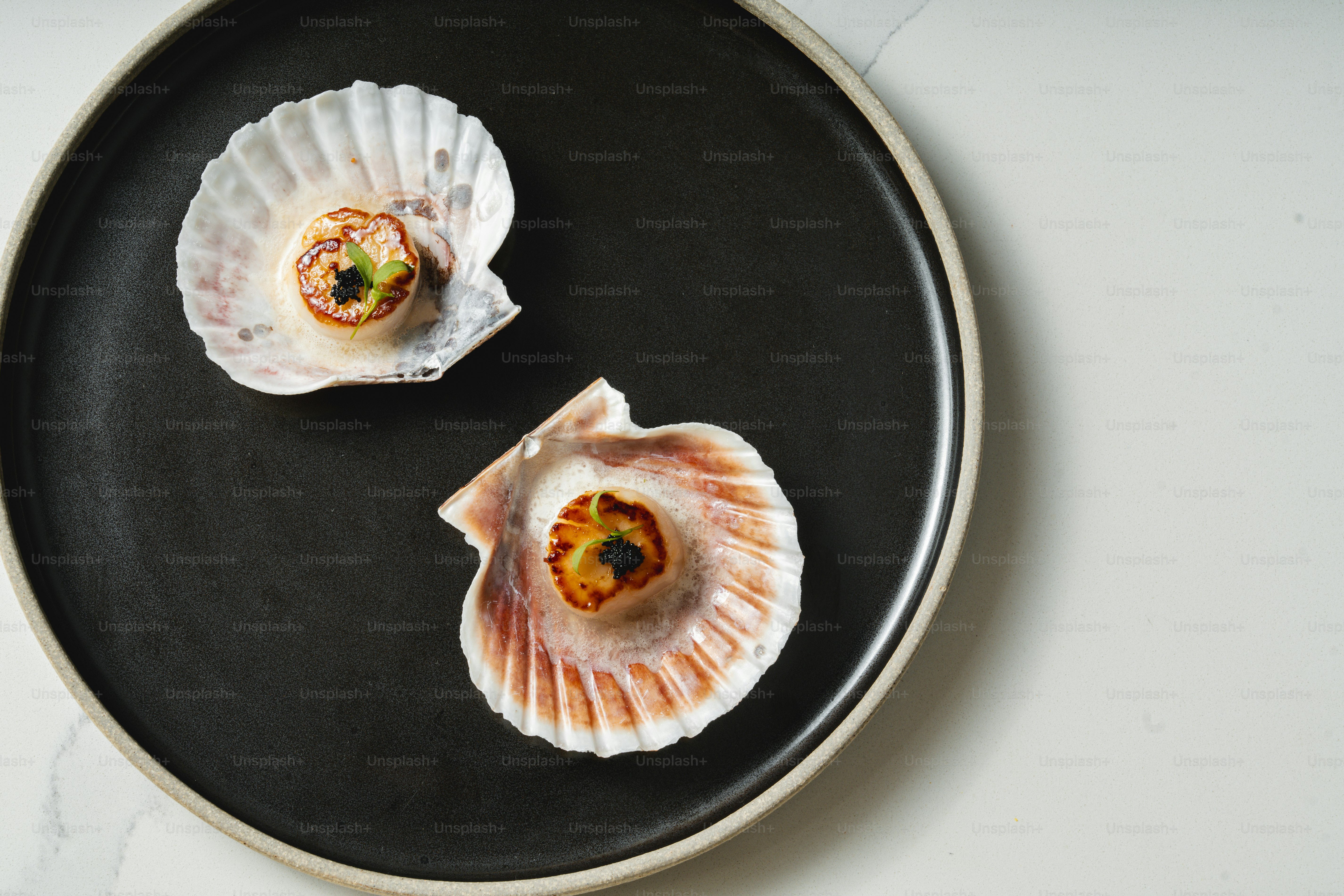 Two scallops on a black plate on a white table