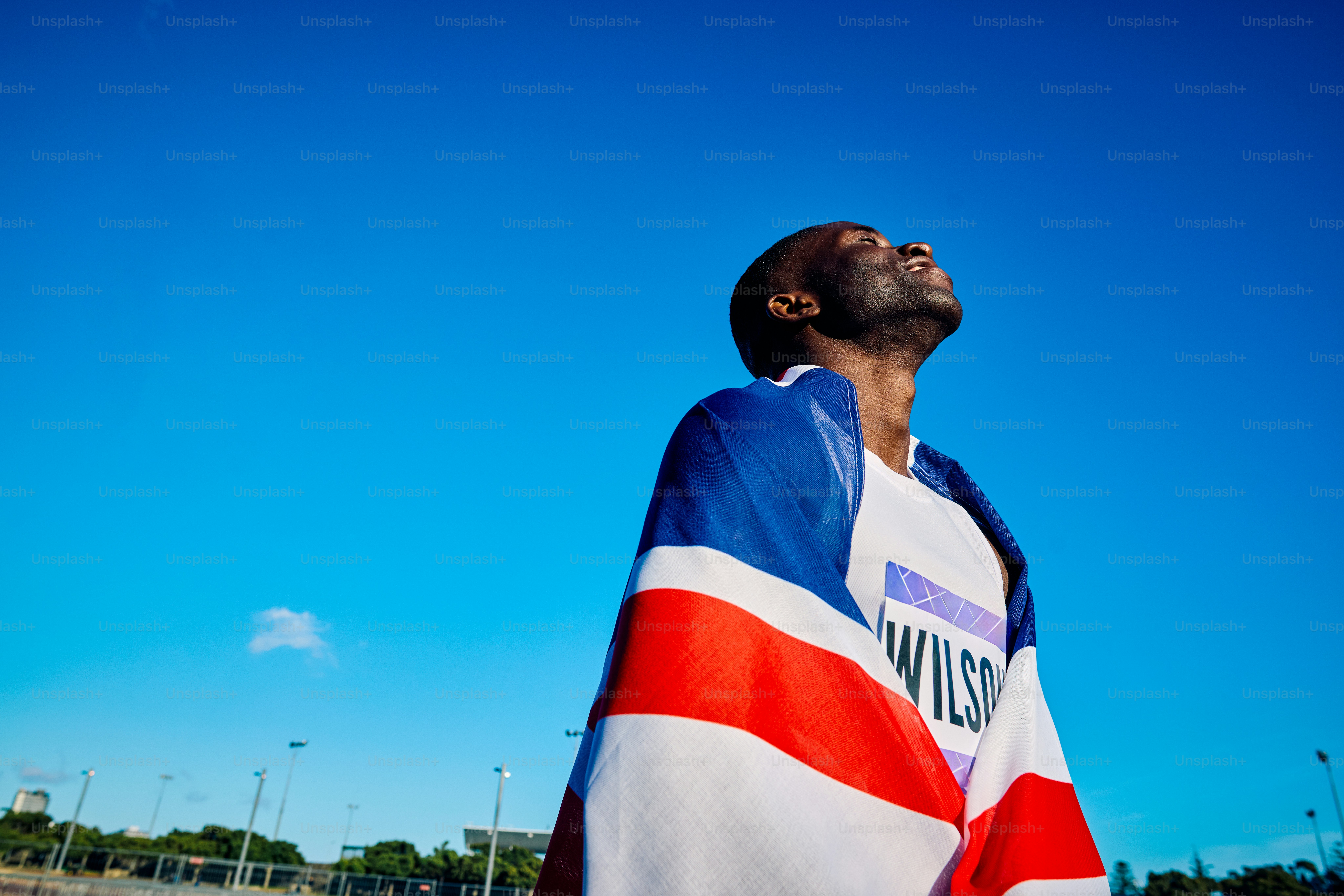 Atlet gembira yang terbungkus bendera Union Jack dalam kemenangan. Potret perayaan luar ruangan dengan langit biru. Konsep kemenangan dan kebanggaan nasional.