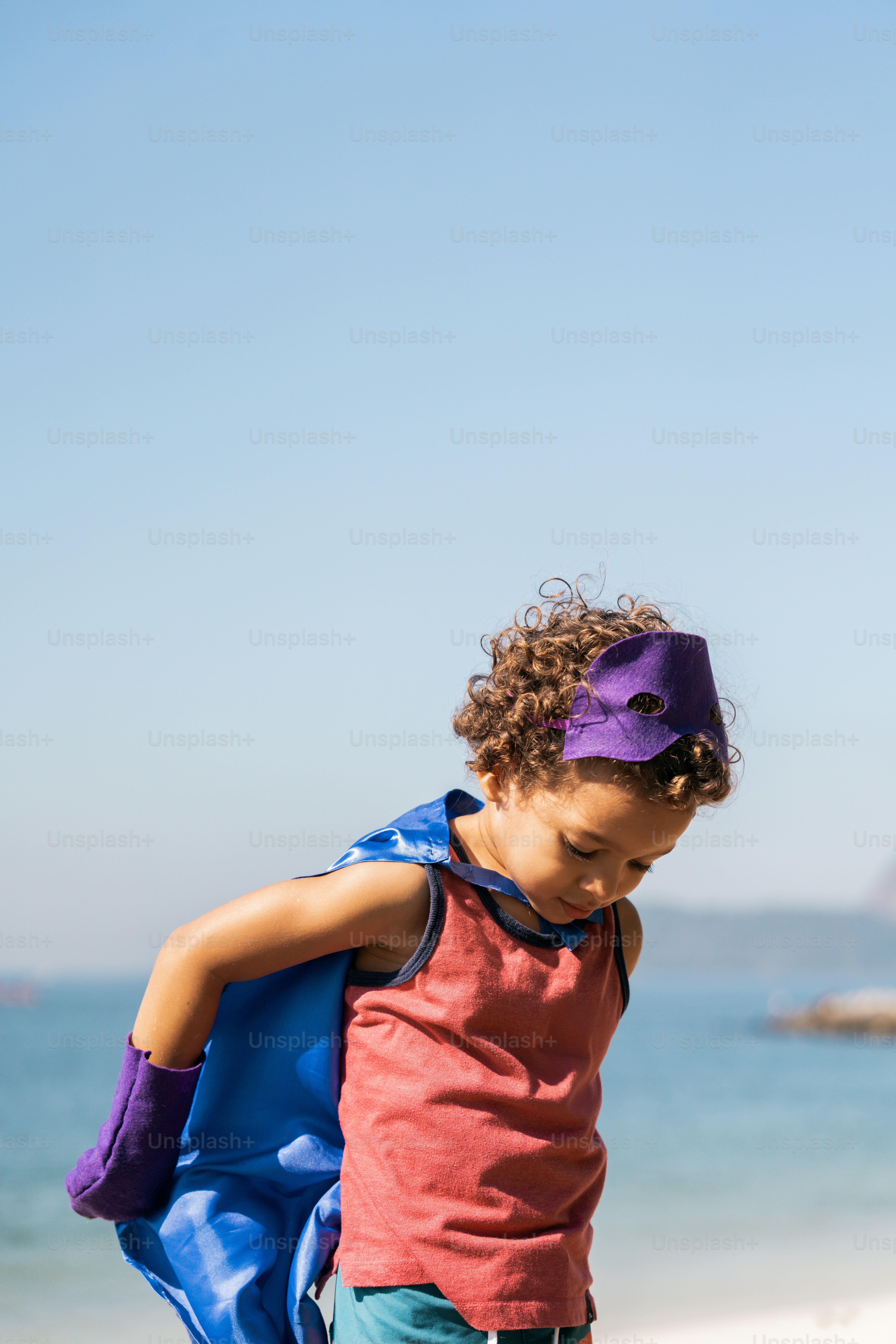 A young boy wearing a purple mask and a blue cape on the beach