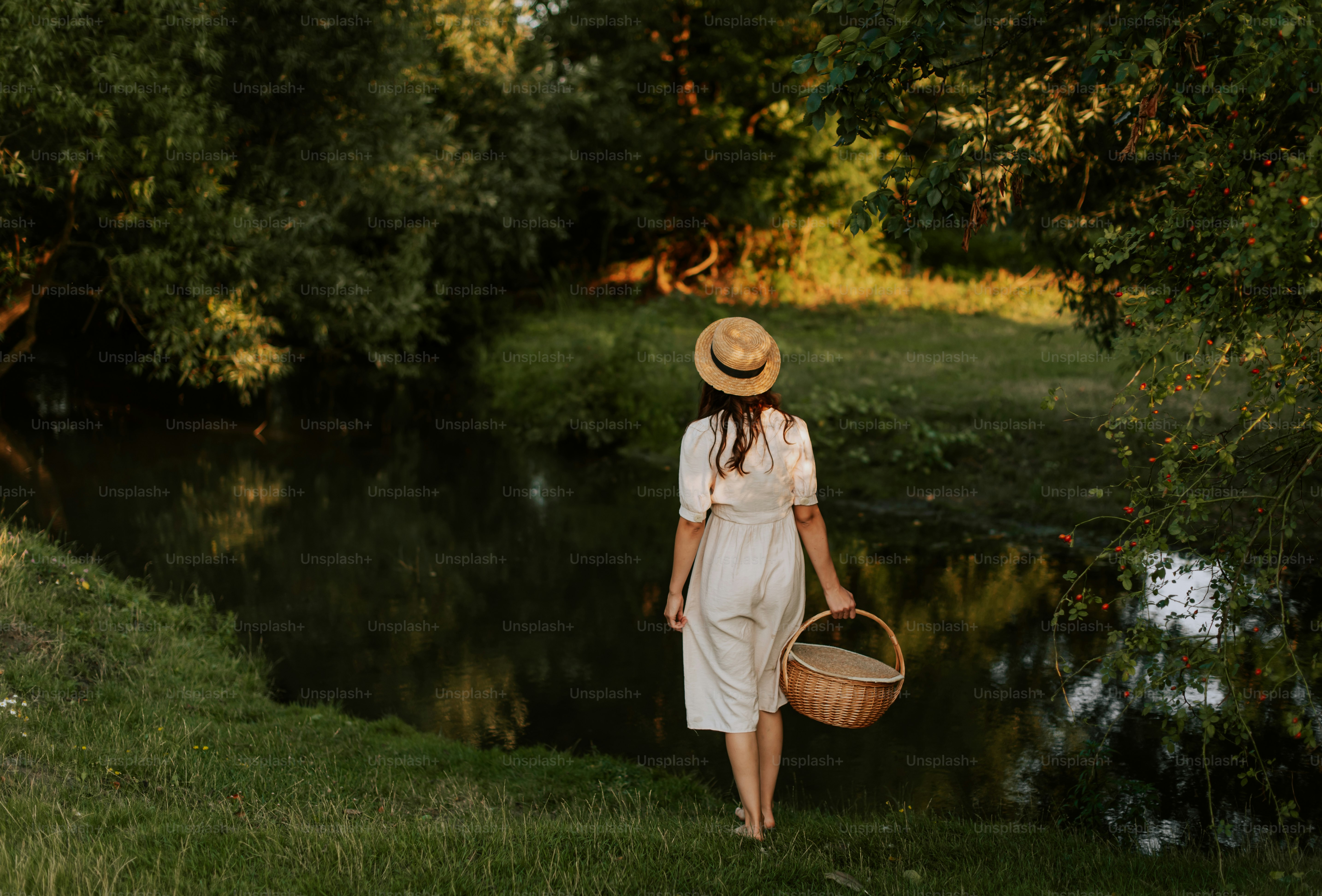 白いワンピースと麦わら帽子をかぶった女性が川沿いを歩いています