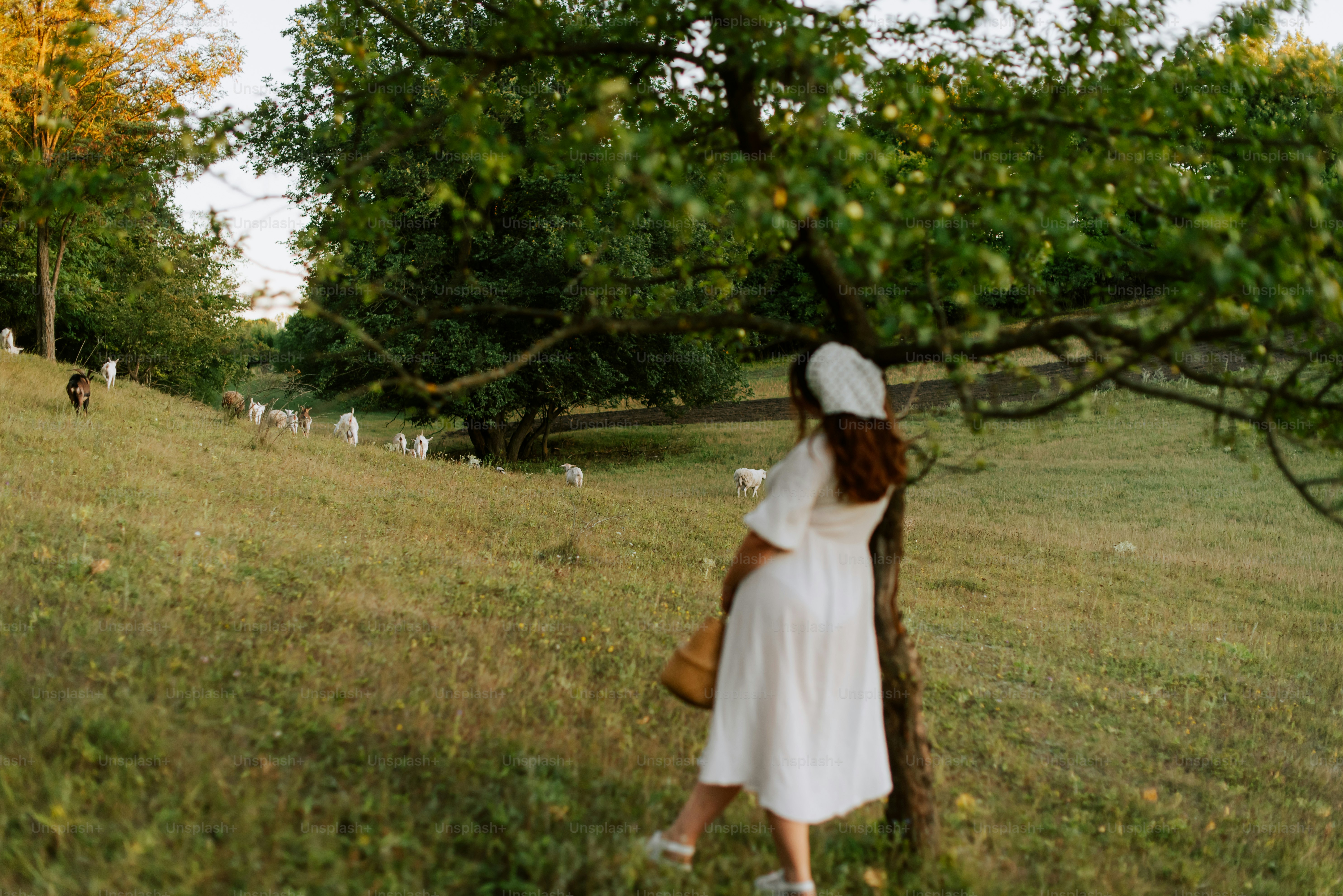 A woman in a white dress and a white hat is standing by a tree