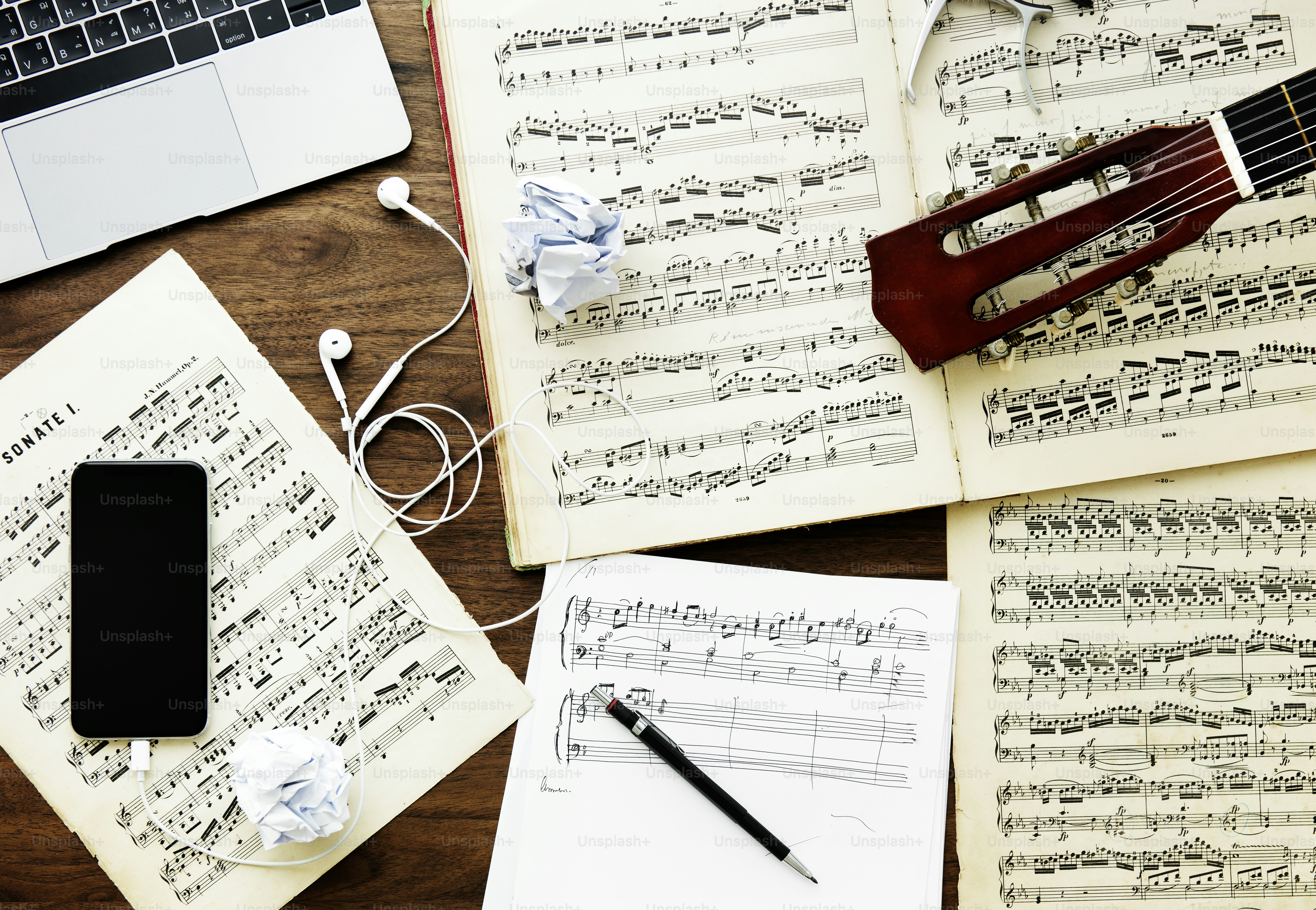 A laptop, smartphone, guitar headstock, and sheet music on a wooden desk