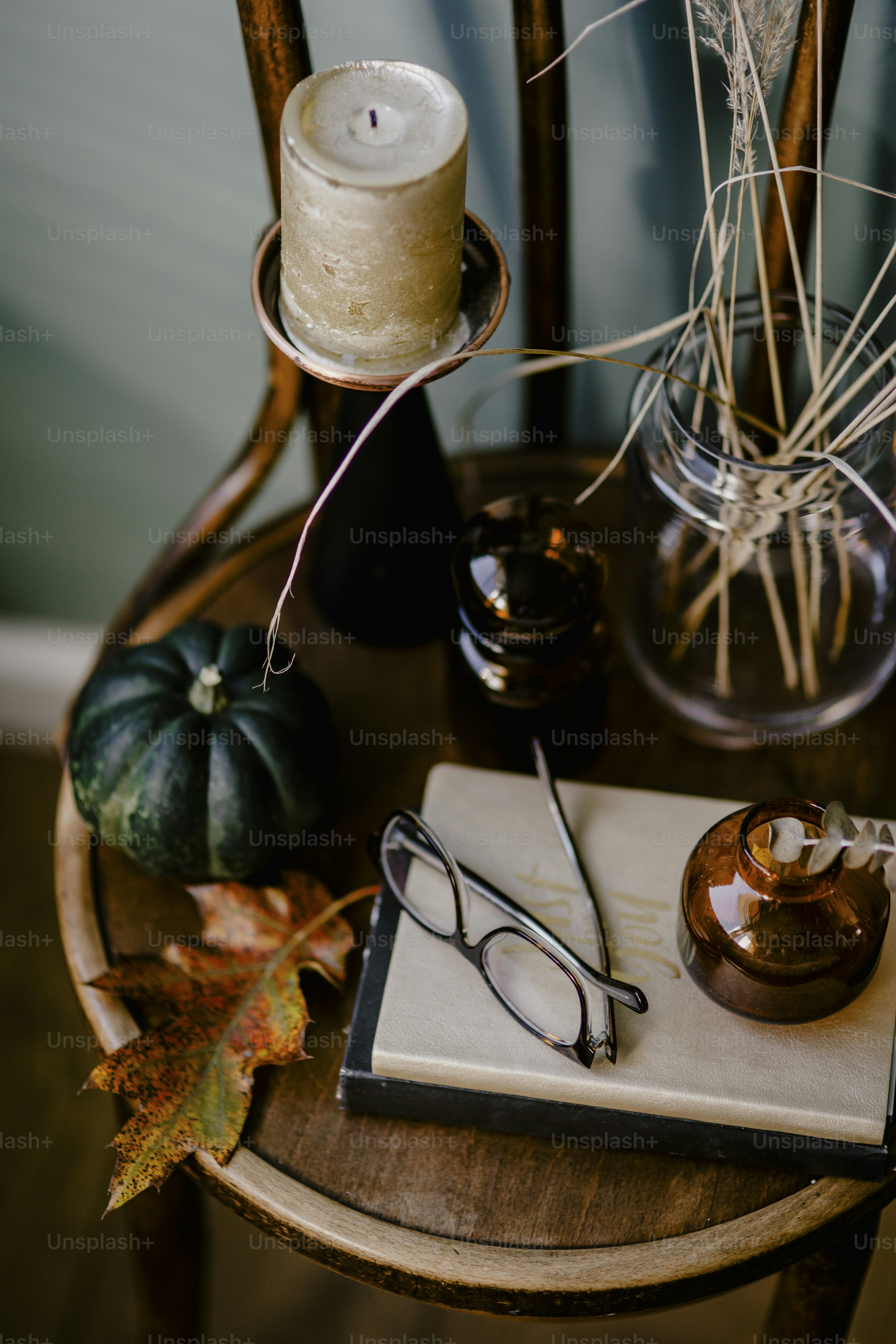 A wooden table decorated with an autumn theme featuring an aromatic candle, glasses, dried flowers, an oak leaf, and a green pumpkin