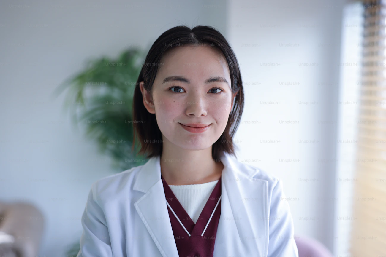 A young Asian female doctor in a white coat looking directly at camera in a clinical office setting