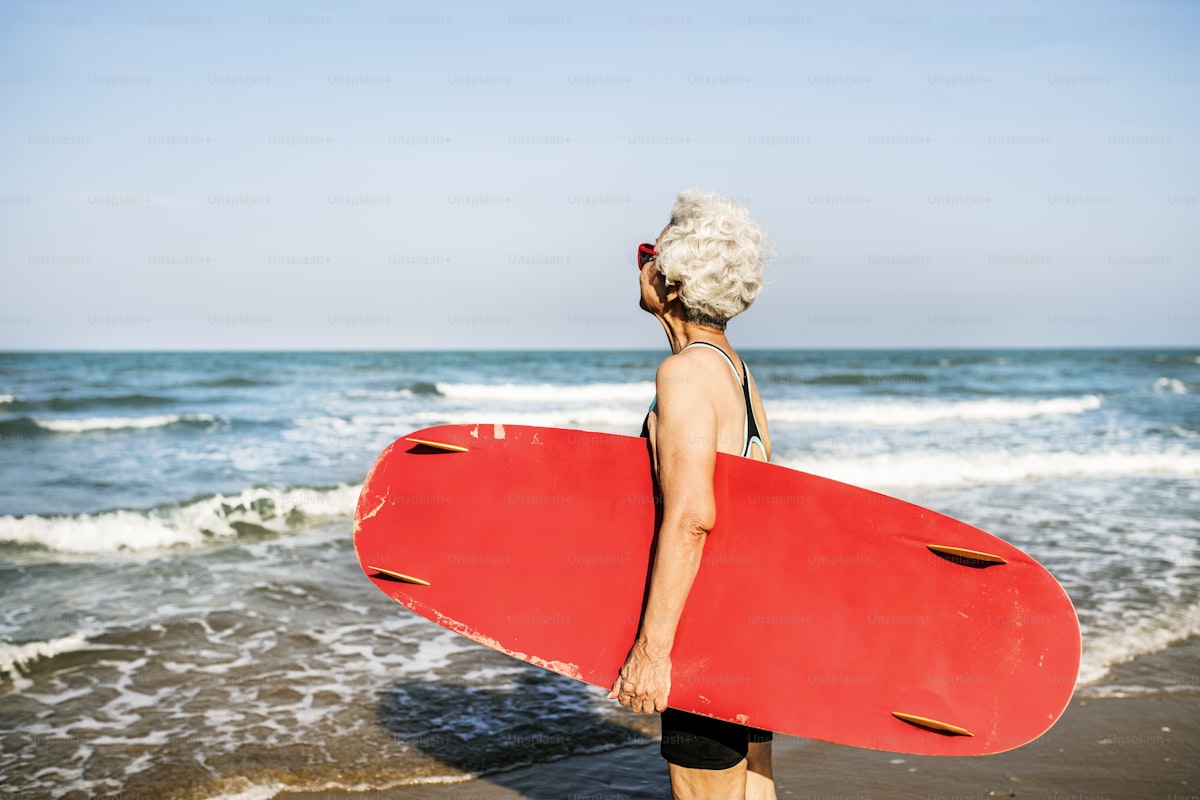 Silver-haired woman carrying a red surfboard at the beach — living her third act
