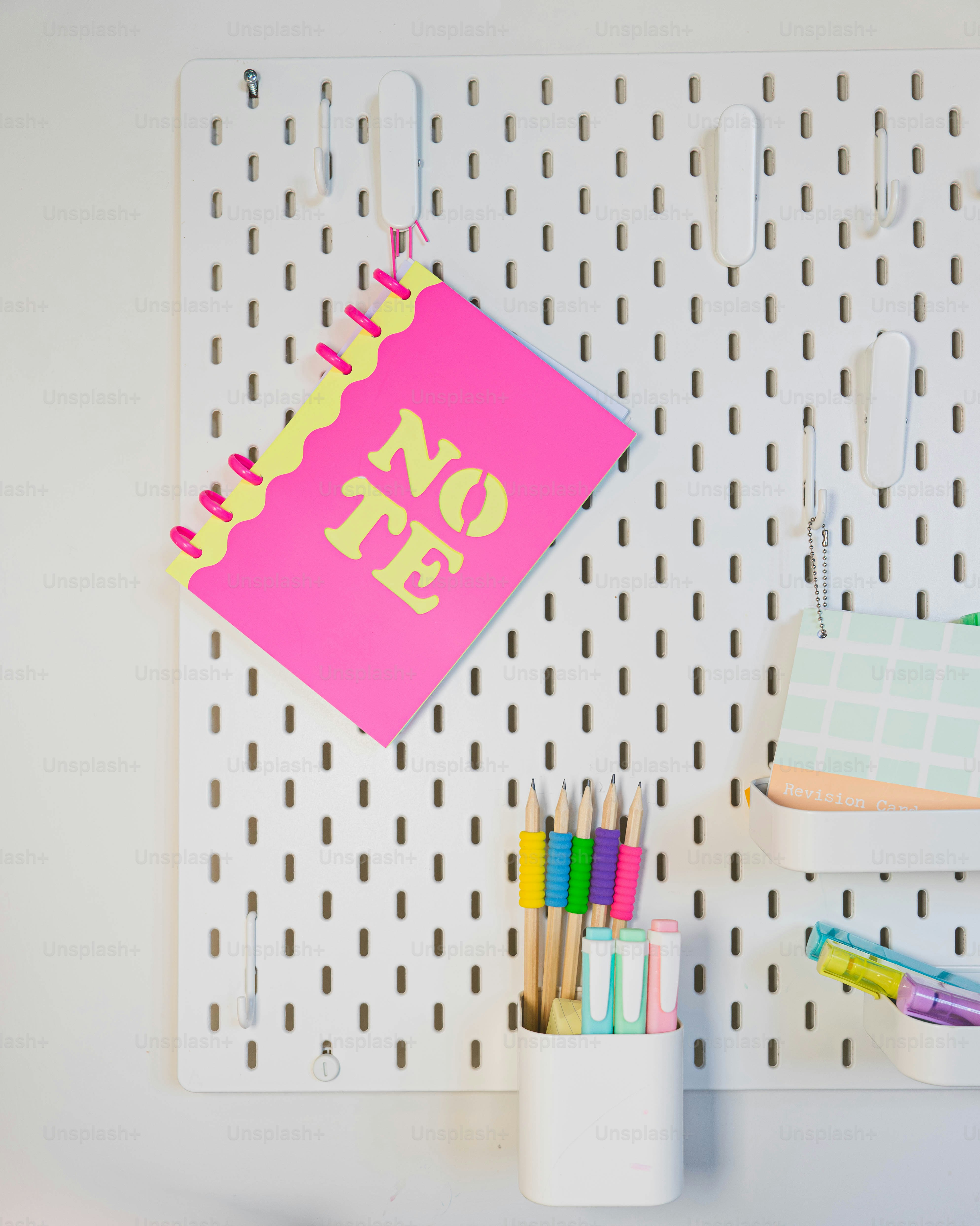 A white pegboard with a pink notepad and some pens and pencils