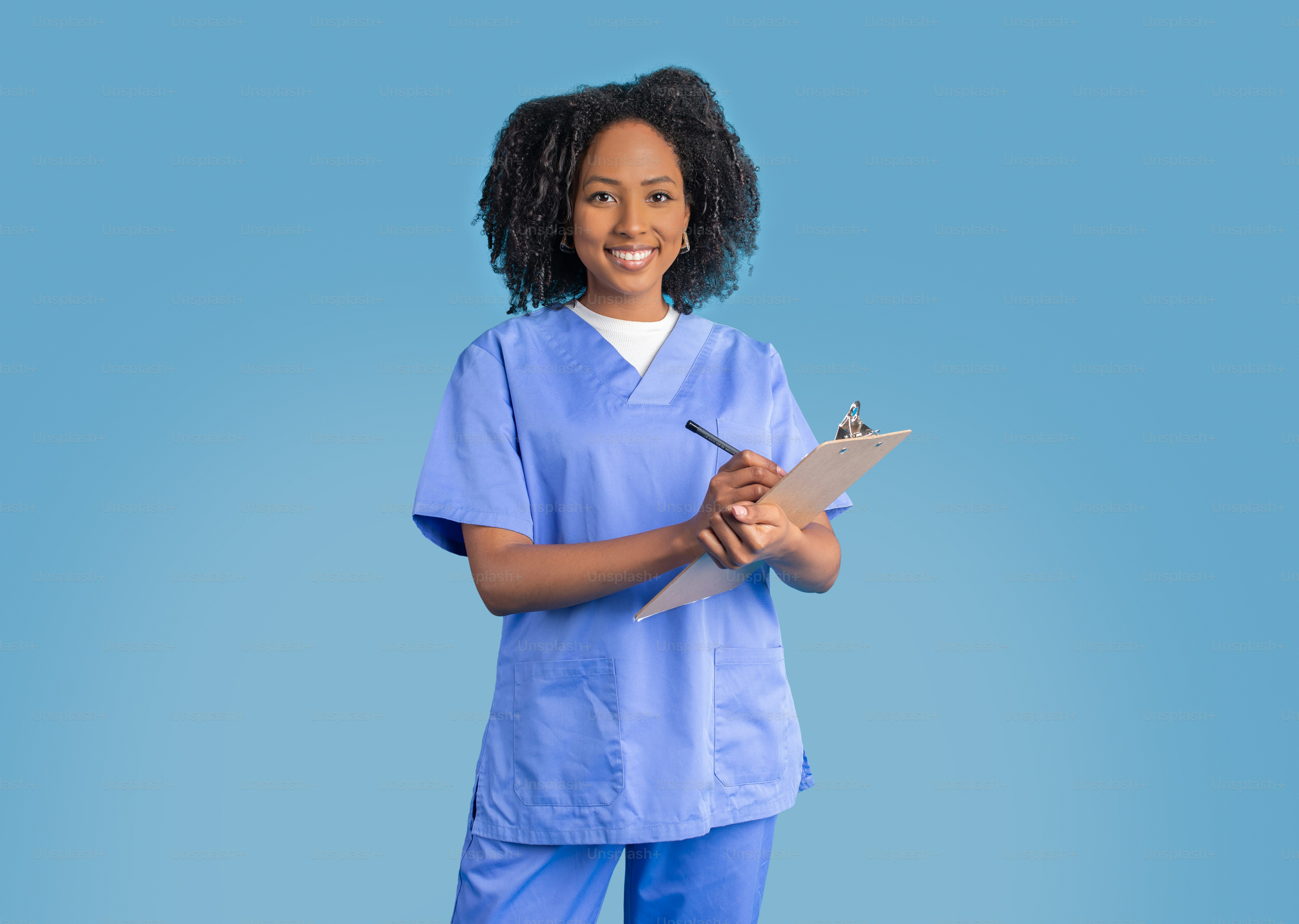 Cheerful young african american curly woman doctor or nurse in blue uniform making notes on tablet, isolated on blue background, studio. Treatment and exam, medicine service, health care