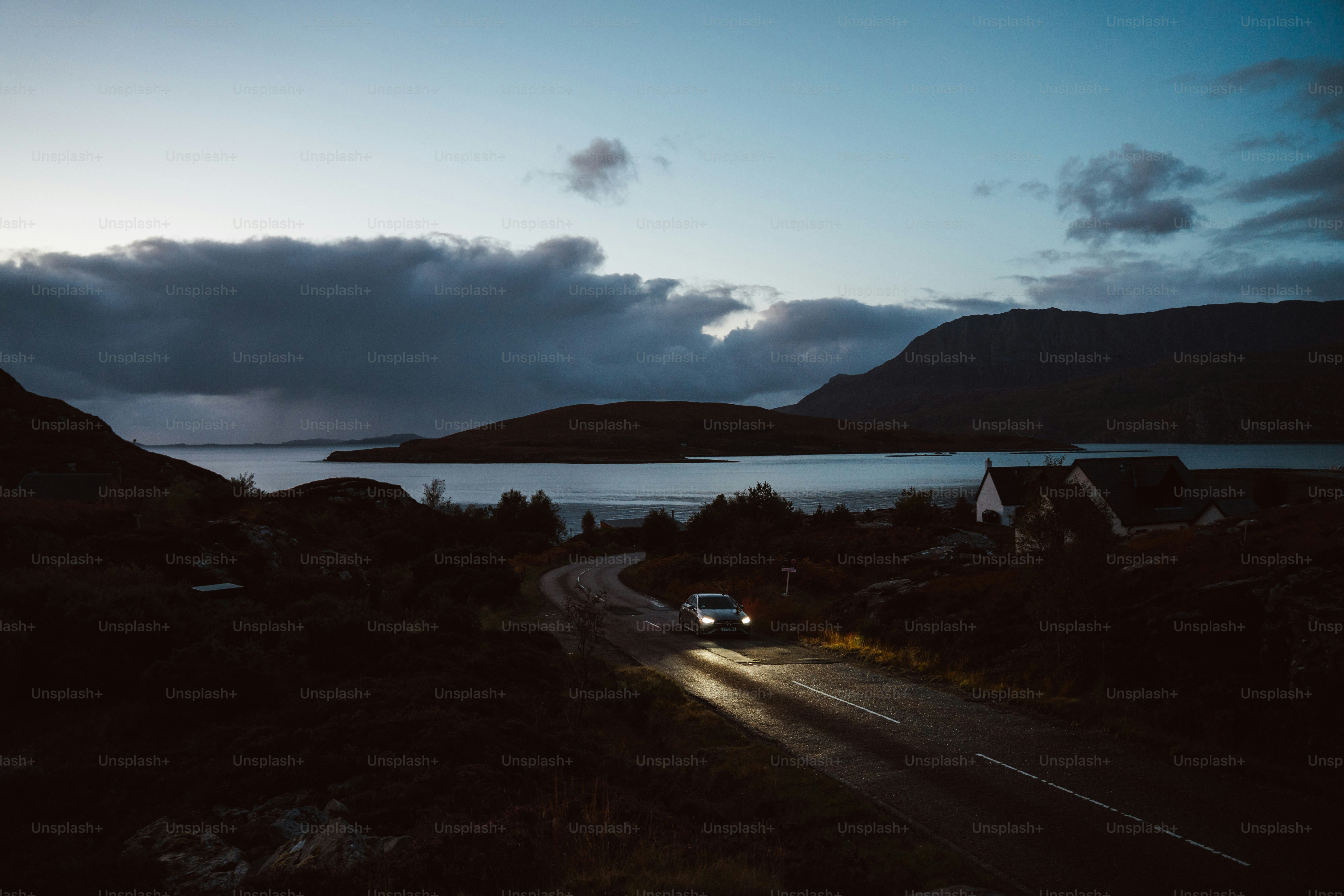 A winding coastal road at dusk — a lone car tracing silver light along cliffs that fall into the sea, the horizon painted in shades of gold and violet.