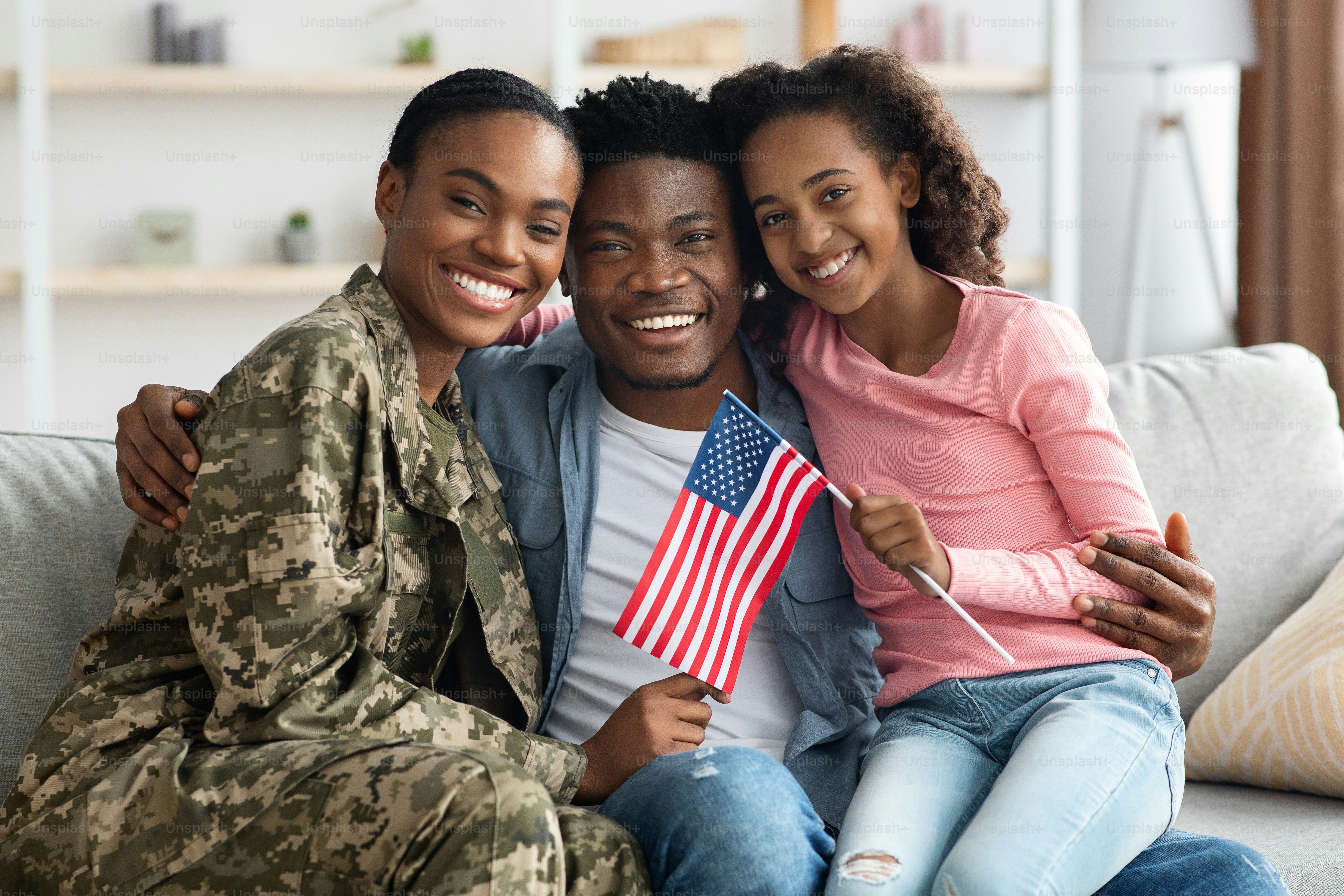 Schöne schwarze Familie, die Wiedersehen zu Hause feiert, glücklicher Vater und Tochter, die ihre Mutter Soldatin begrüßen, auf der Couch sitzend, umarmend und lächelnd in die Kamera, Flagge der USA halten