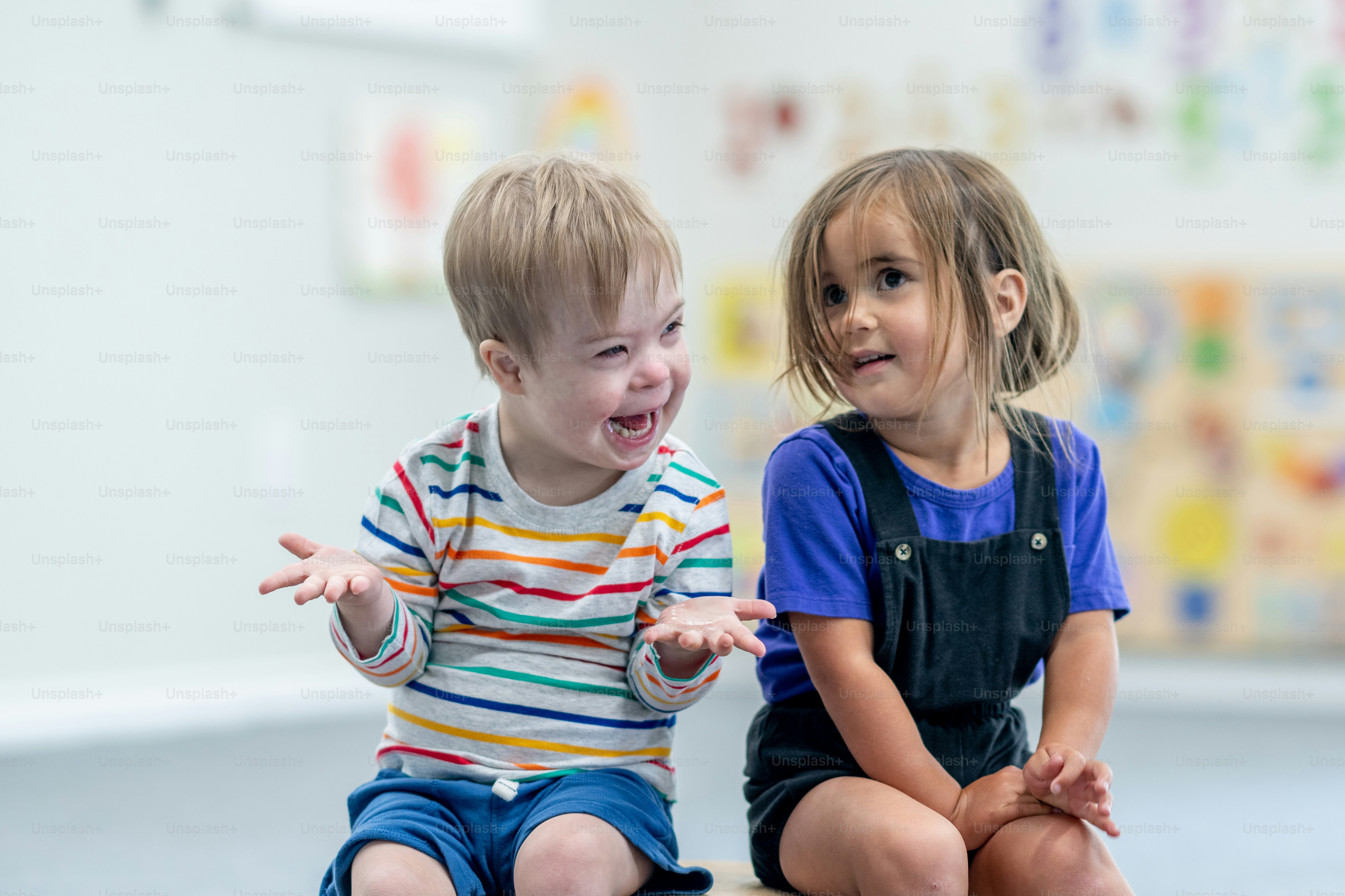 Two preschool aged children sit on a small wooden bridge as they pose together for a portrait.  They are both dressed casually and are smiling.