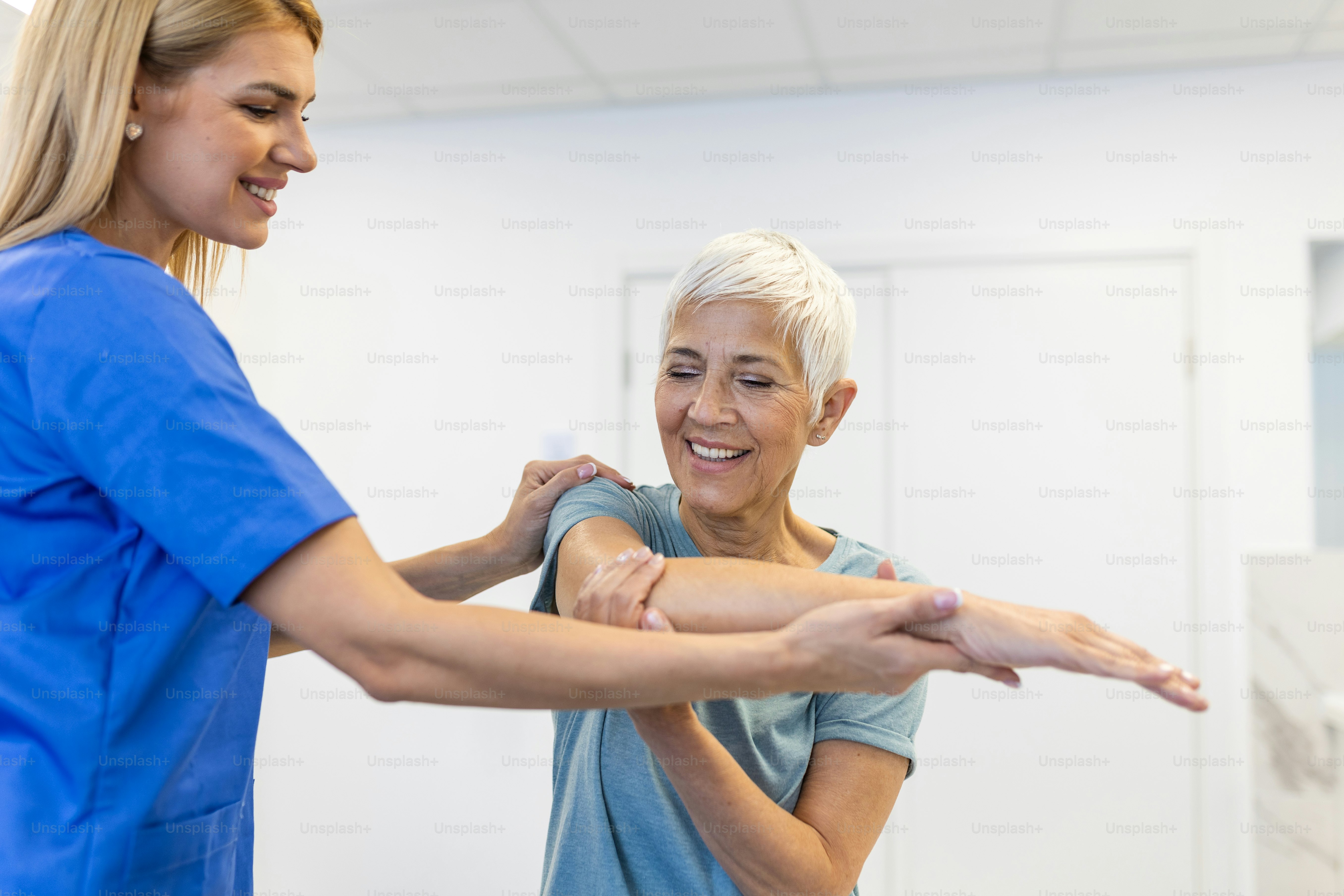 Happy senior woman doing exercise with physiotherapist. Old retired lady doing stretching arms at clinic with the help of a personal trainer during a rehabilitation session.