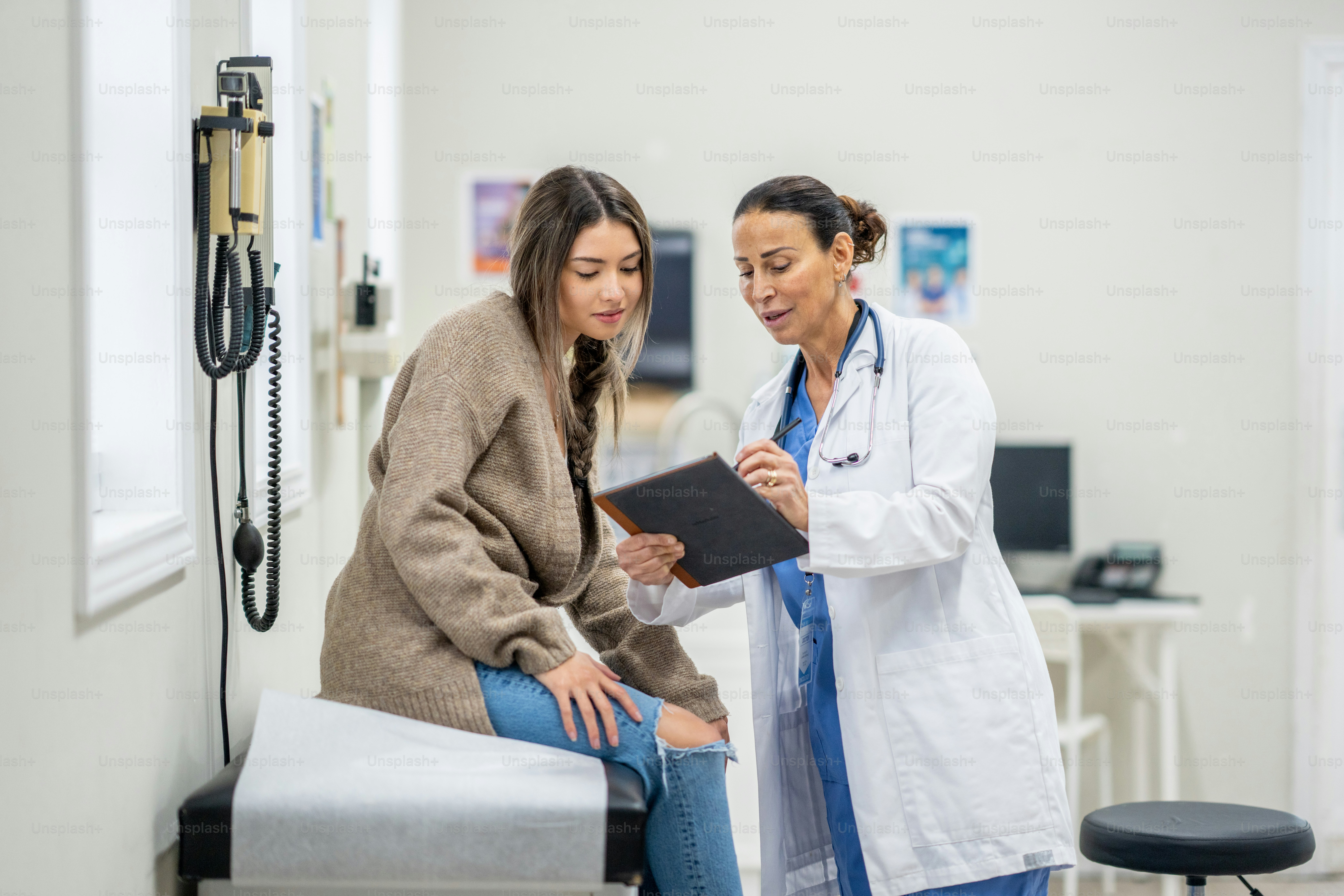 A young female teenager sits up on an exam table during a routine medical appointment with her doctor.  She is dressed casually and leaning in to look at her test results as her doctor shares them on a tablet