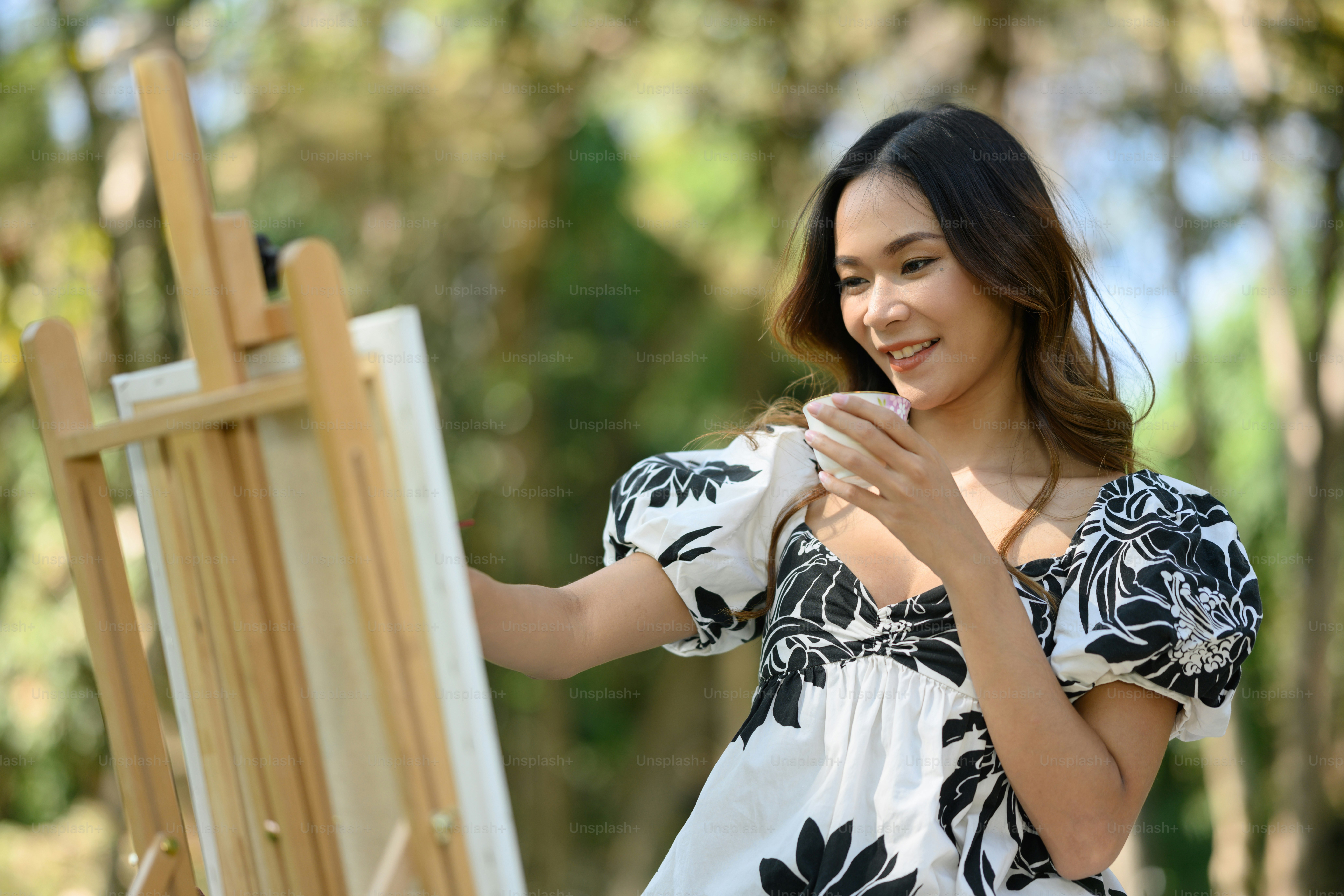 Relaxed young woman holding cup of tea and painting on canvas at the park.
