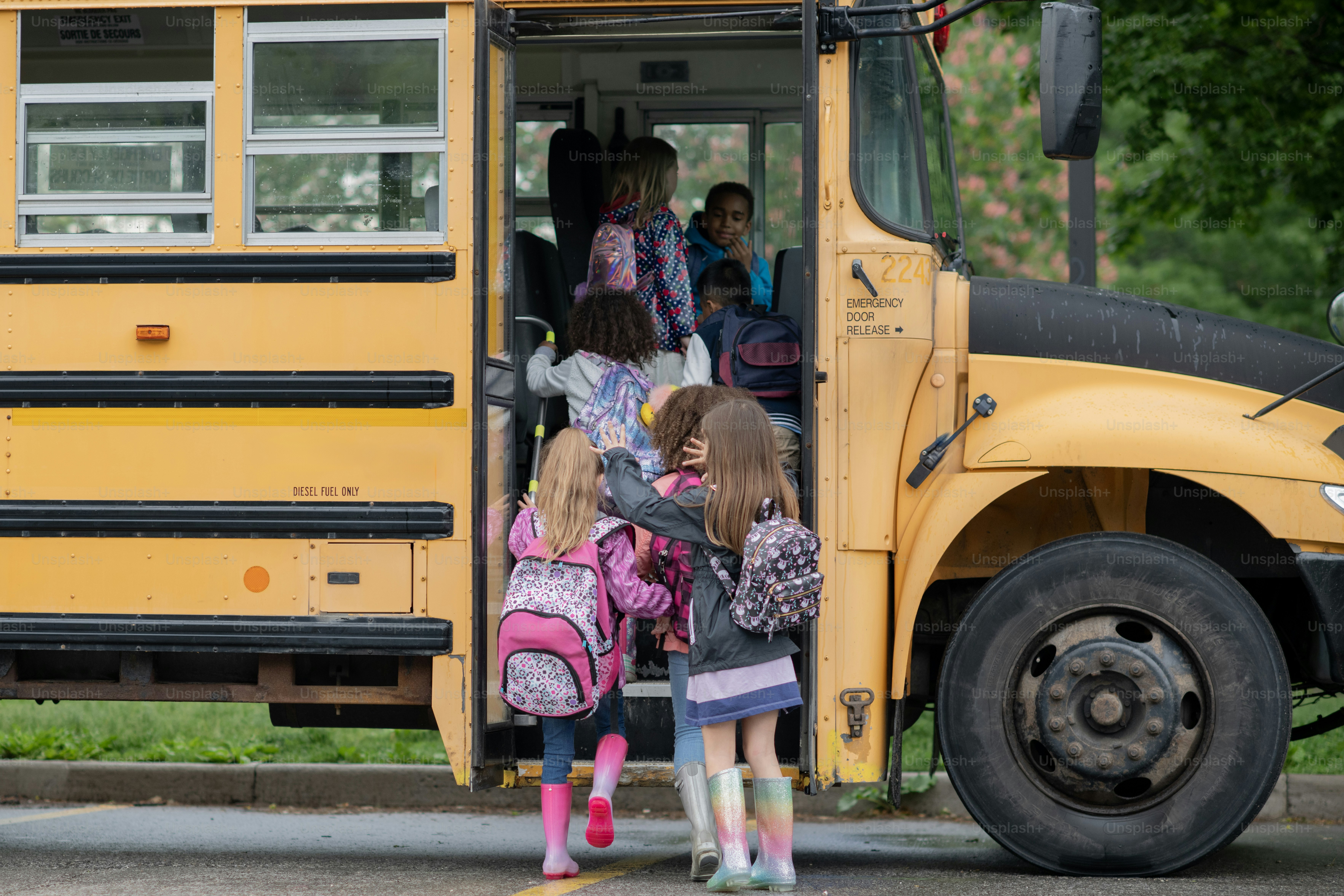 A large group of school aged children are seen boarding the school bus in the morning as they make their way to school.  They are each dressed casually and have backpacks on.