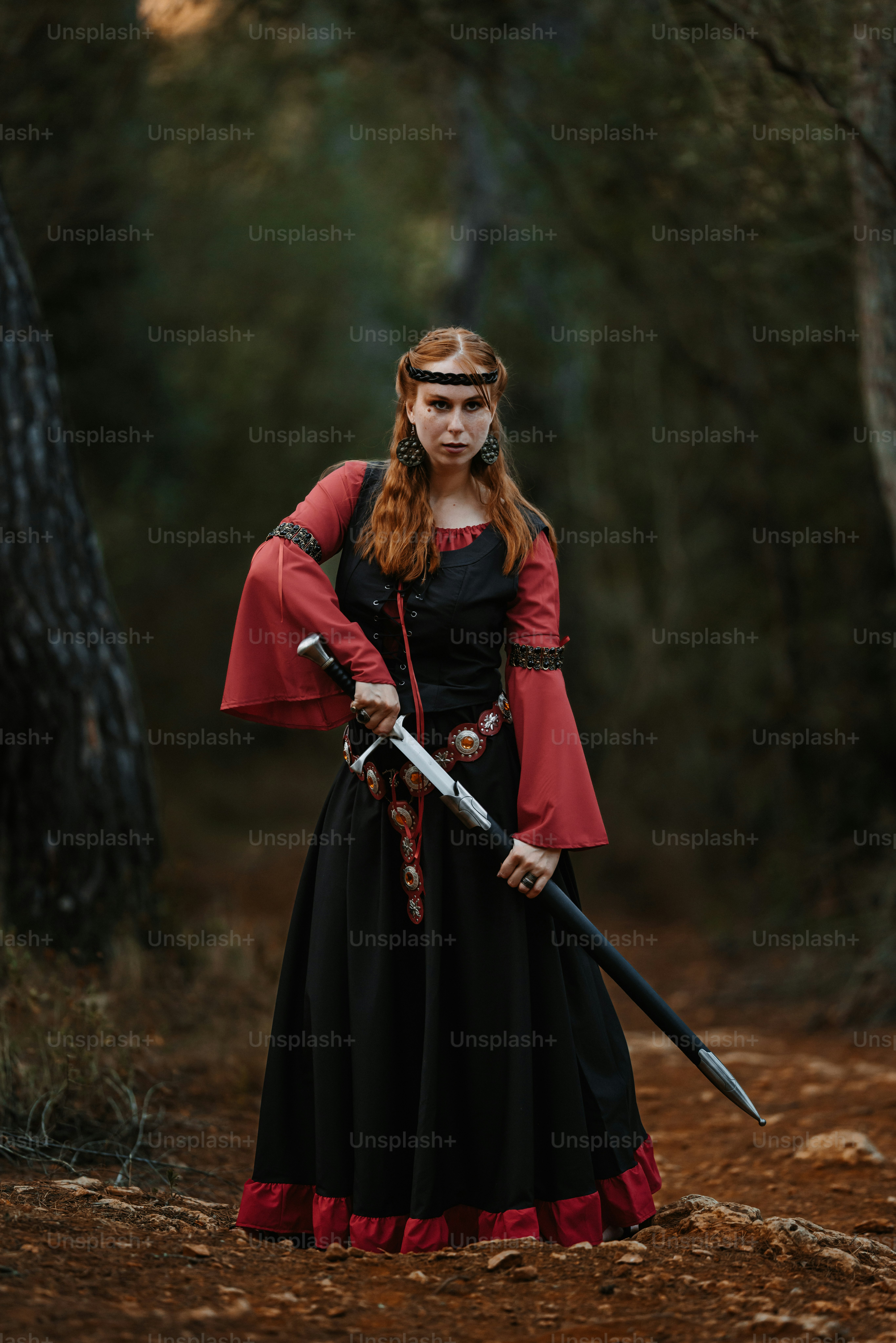 Young red-haired medieval fantasy sorceress stands in the forest looking directly into the camera and with a determined gaze she draws her sword. Creative dramatic color retouching and grain added. Part of a series.