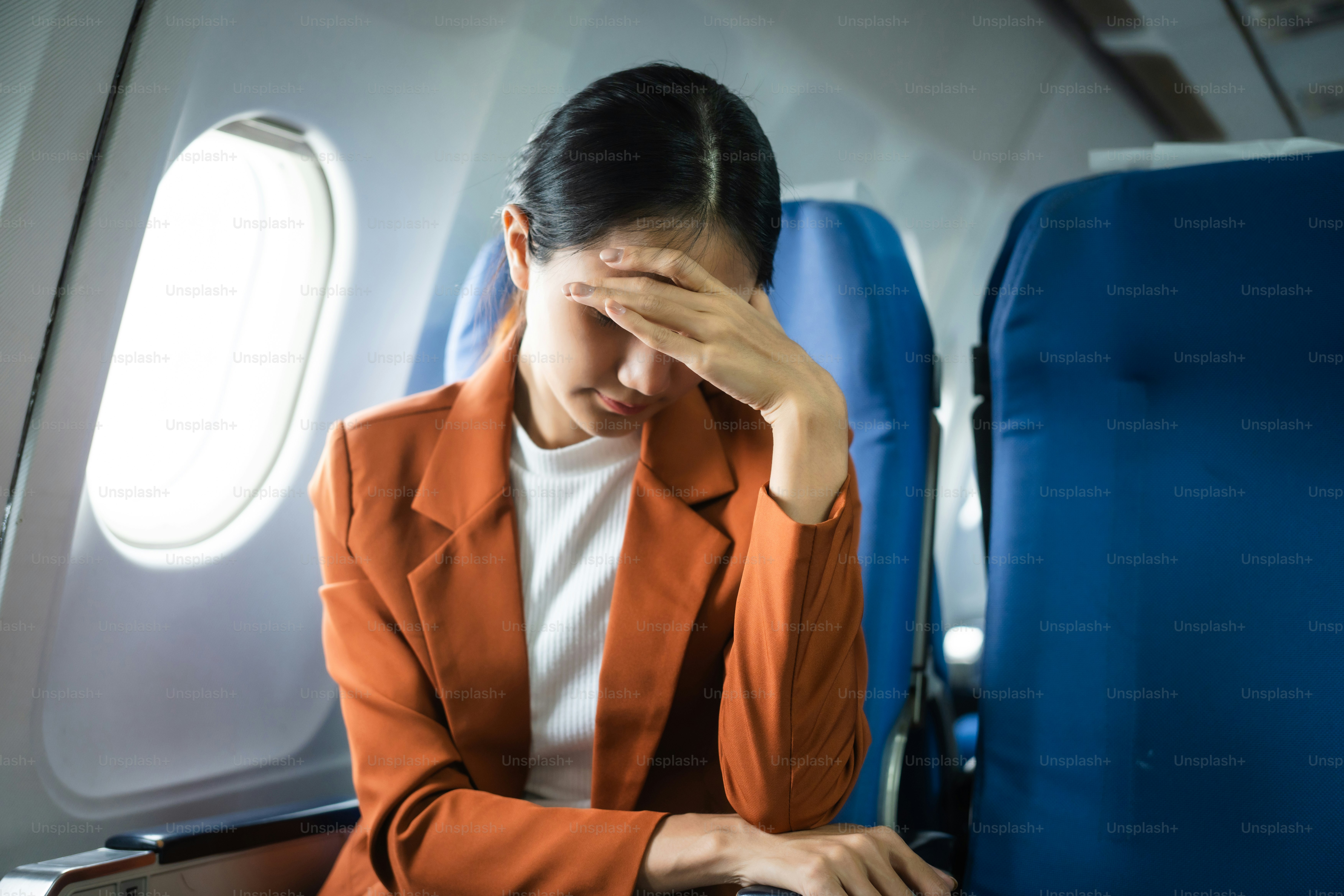 A young Asian woman, an airplane passenger, sits by the window seat, experiencing nausea and dizziness during the flight, which adds to her travel discomfort.