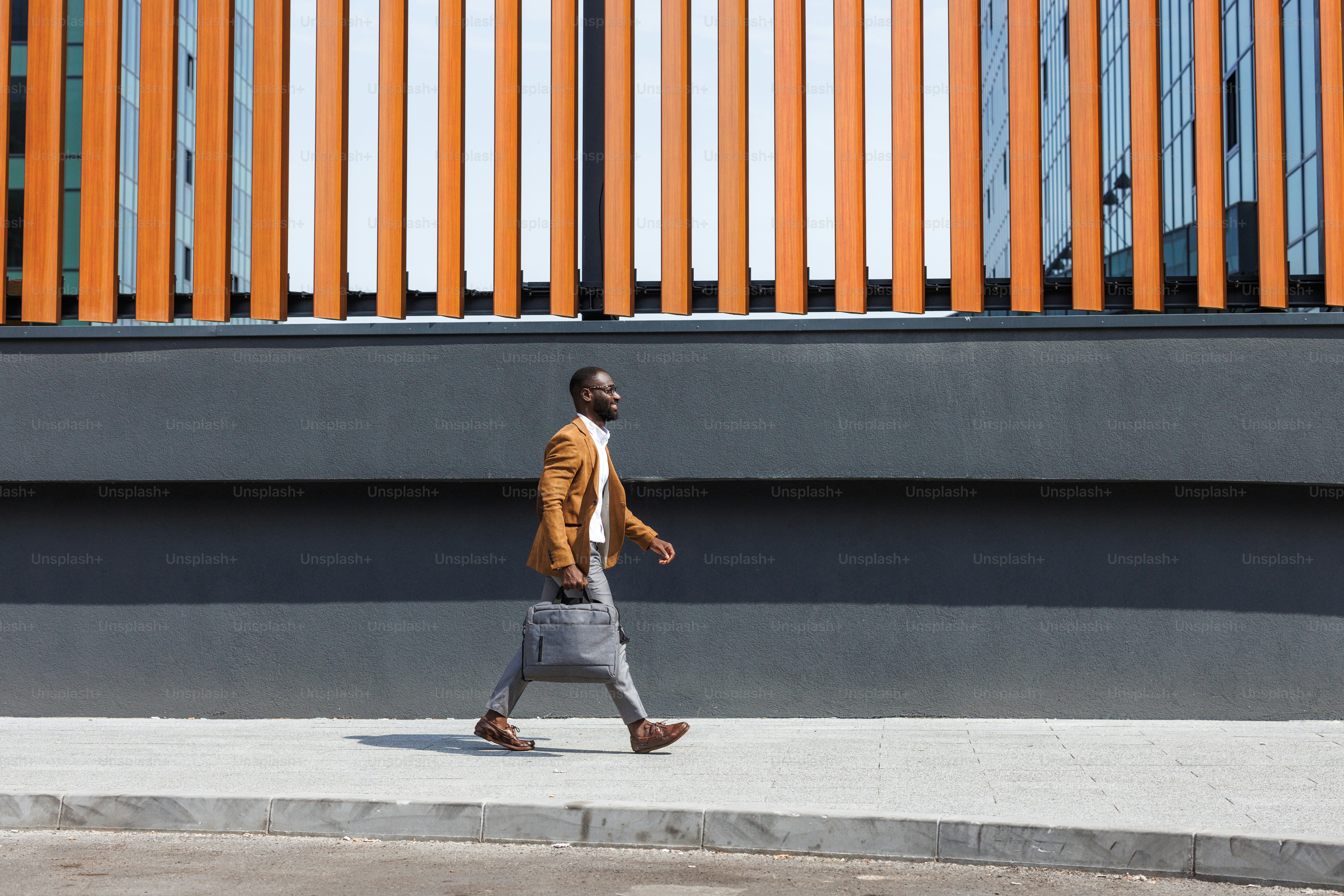 Side view of a confident businessman in a stylish outfit walking outdoors on a sunny day, exuding professionalism and determination in a modern urban environment.