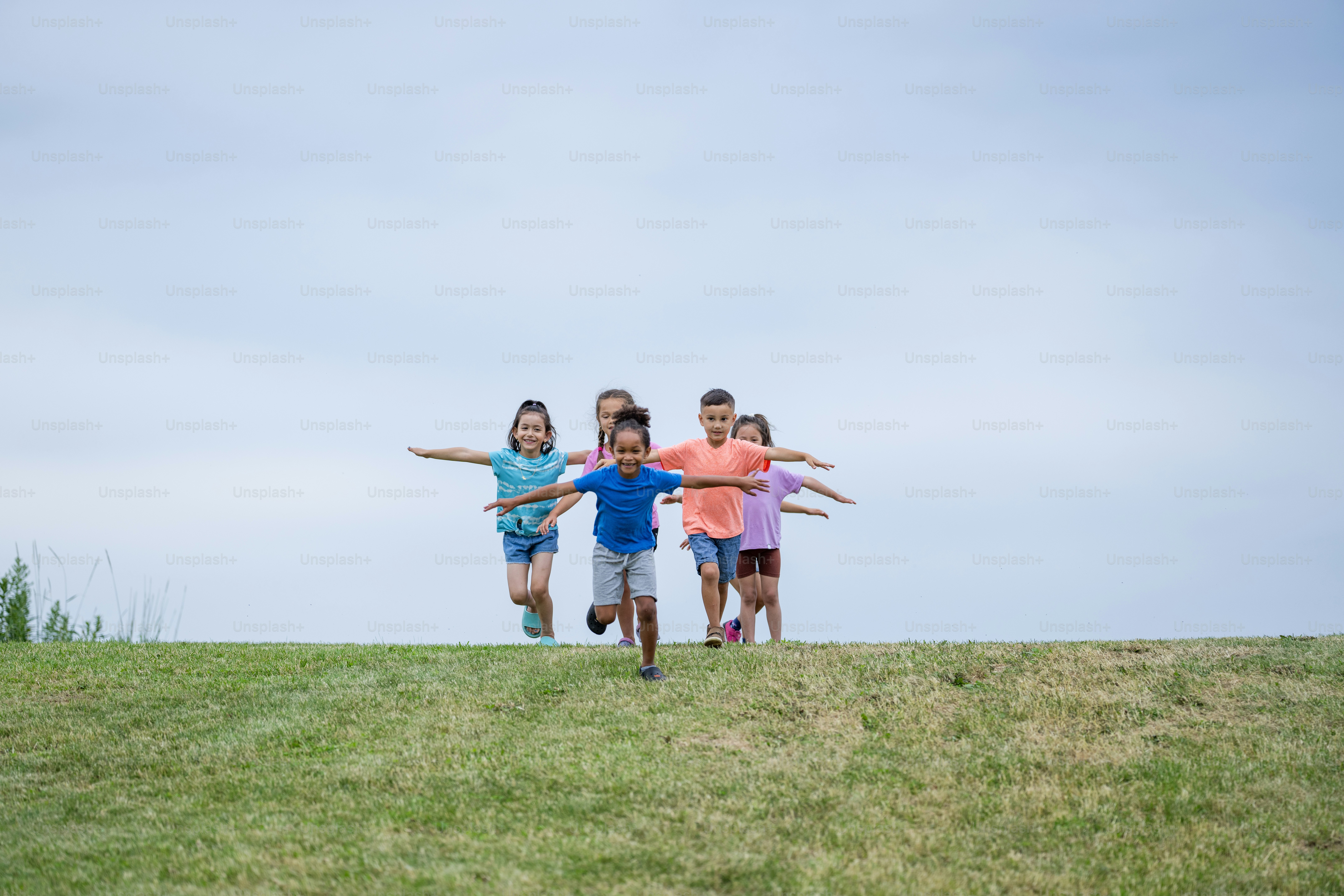 A small group of young children are seen running down a grassy hill with their arms out like airplanes as they enjoy the fresh air.  They are each dressed casually and are laughing and smiling.