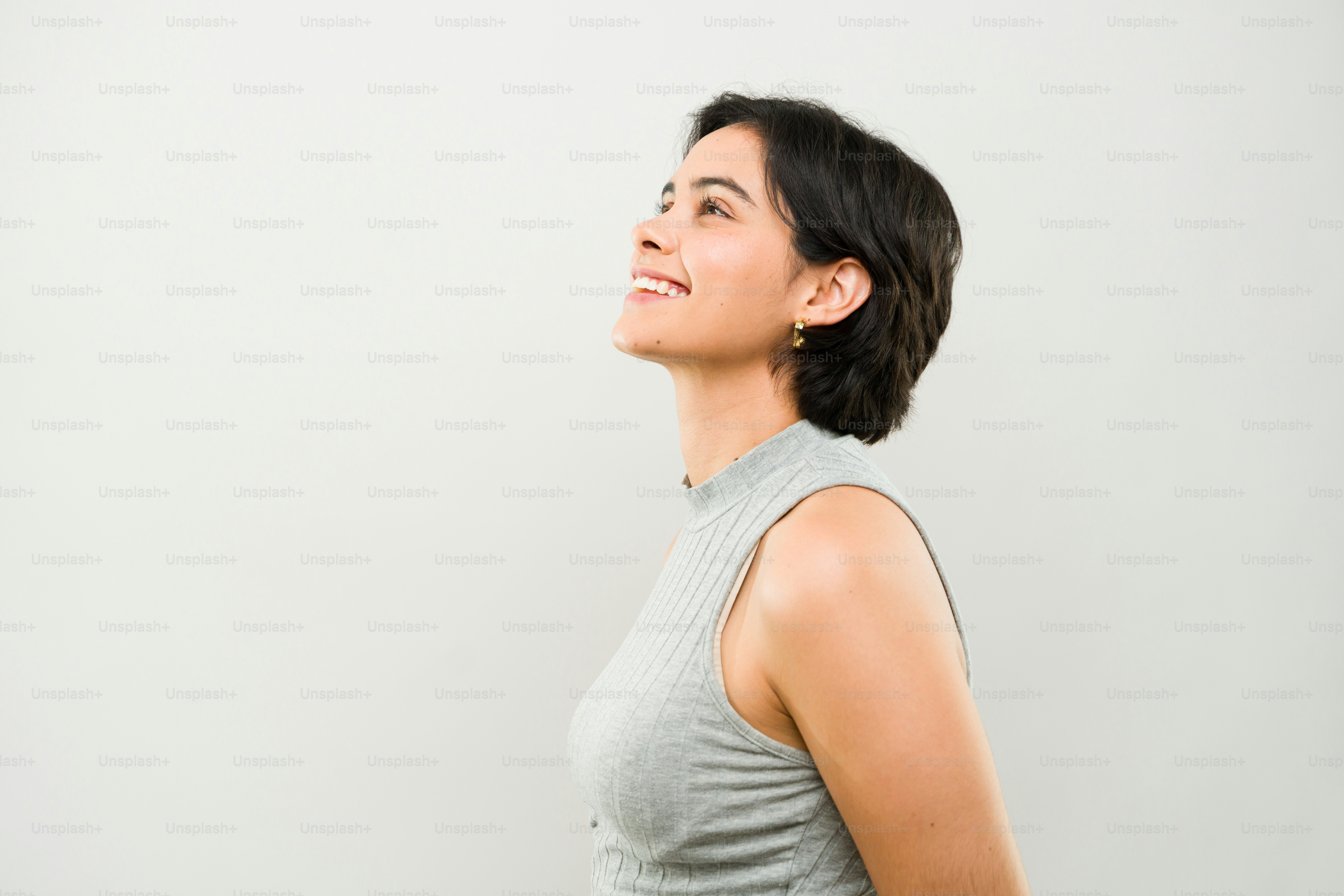 Jeune femme latine aux cheveux courts portant une chemise grise souriant et levant les yeux sur un fond blanc