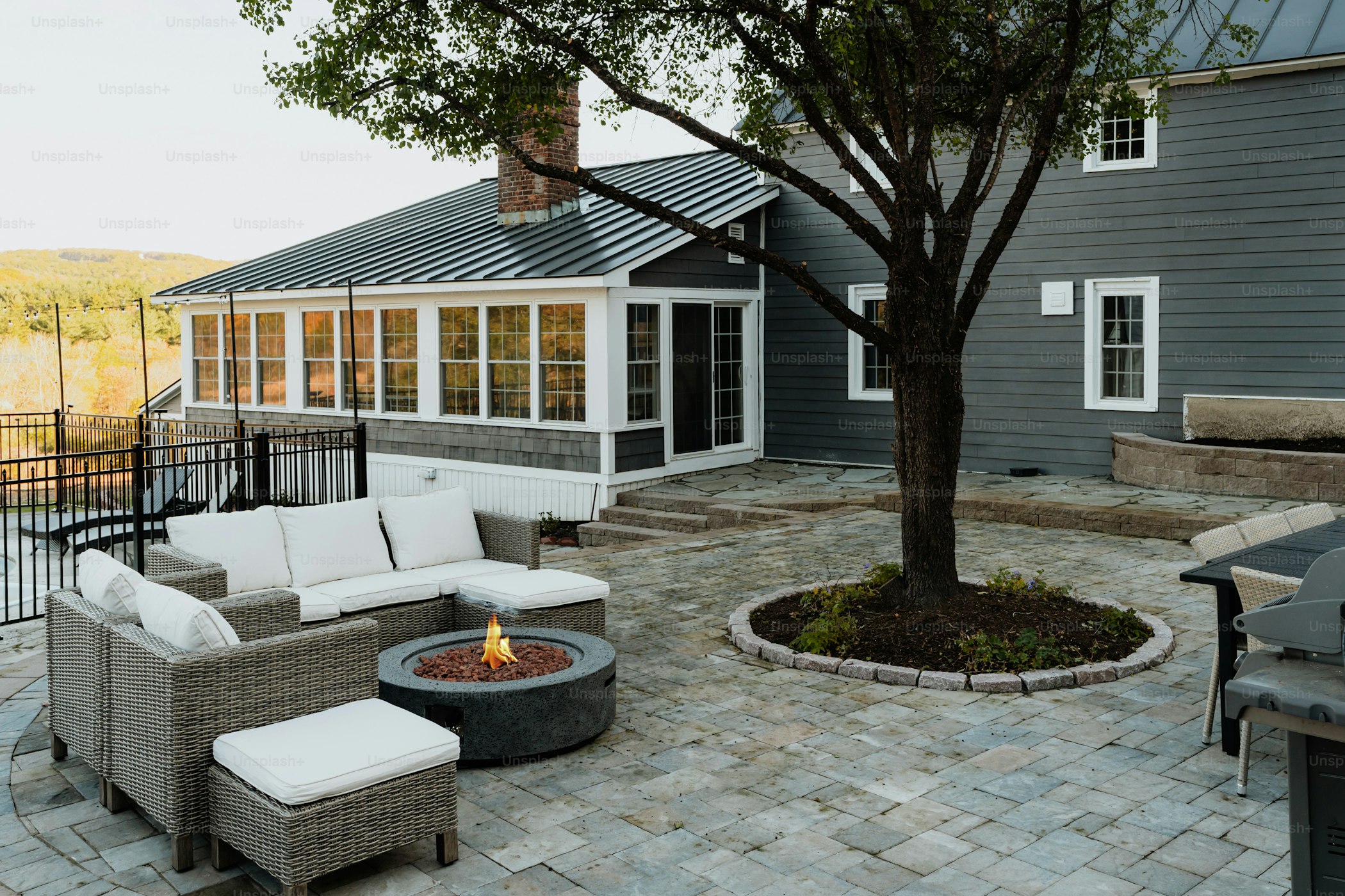 Stone patio with built-in fire pit and Adirondack seating, surrounded by mature trees and evening light